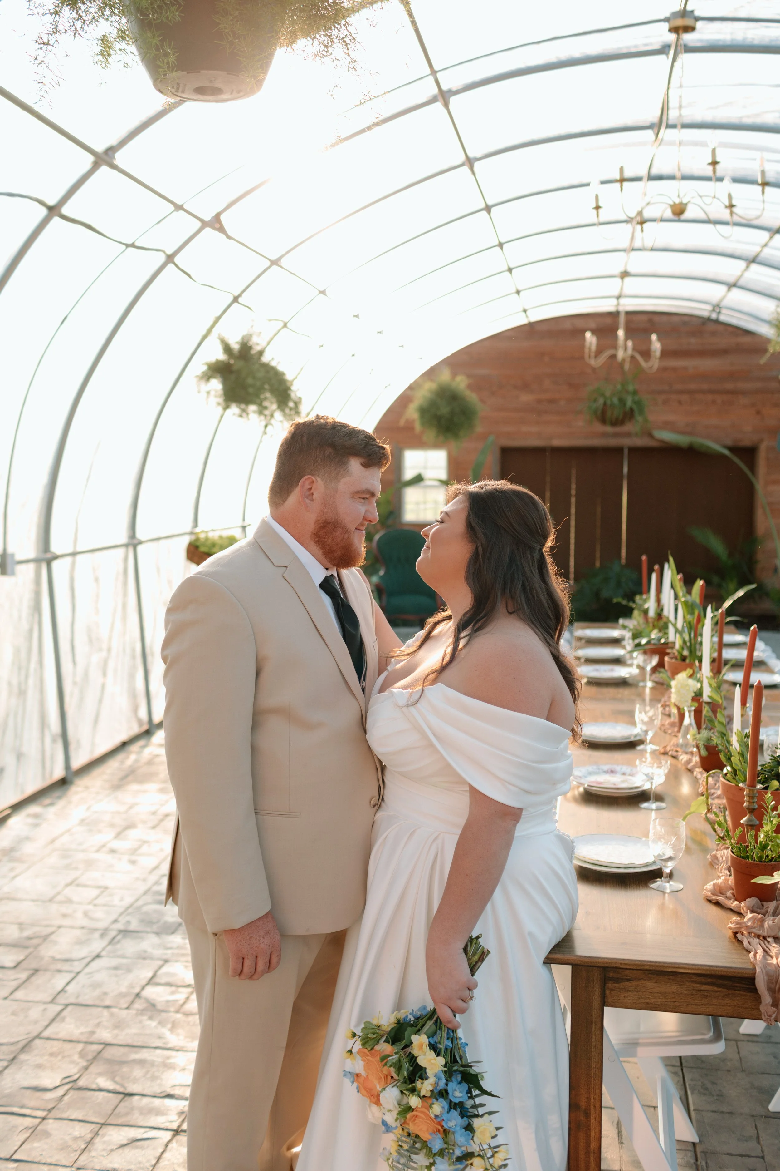 A bride and groom are standing close together in a greenhouse, gazing at each other. The bride is holding a colorful bouquet, and they are surrounded by a long dining table decorated with candles and flowers.