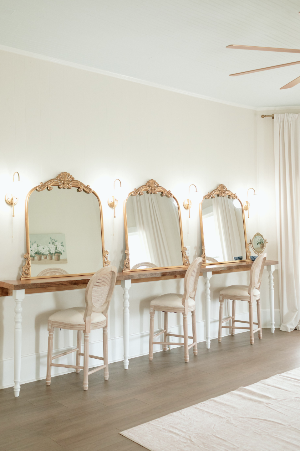 A row of three vintage-style mirrors with ornate gold frames and matching sconces above each mirror, set on a wooden vanity with four white legs and cream-colored chairs with cane backrests, in a bright room with cream walls, white curtains, and a ce