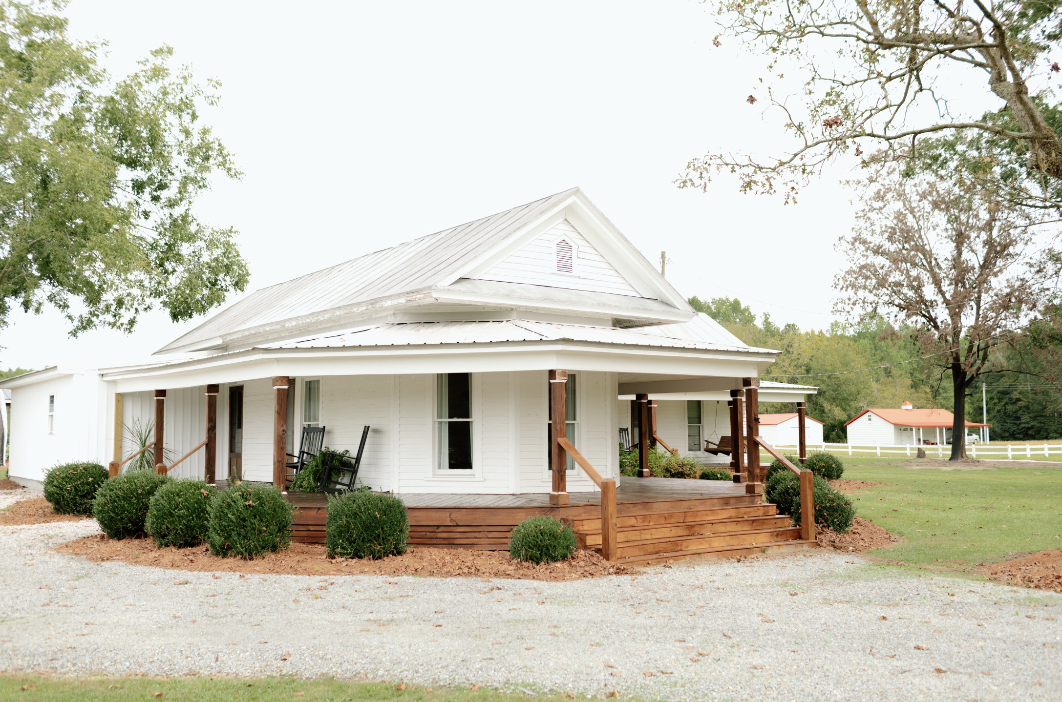 White house with a metal roof, porch with wooden railing, rocking chairs, and surrounding shrubs, trees, and grass.
