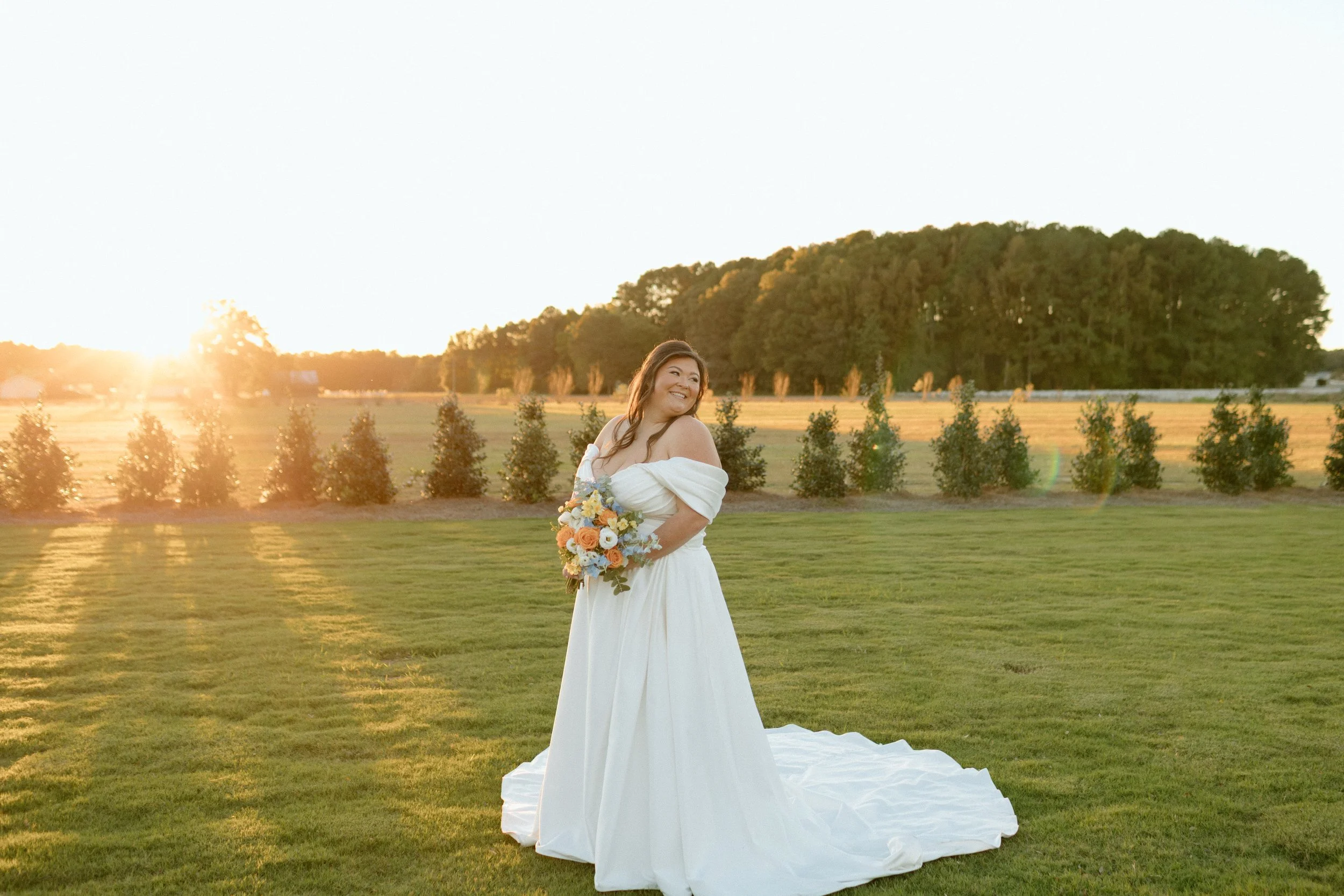 A woman in a white wedding dress holding a bouquet of flowers standing on green grass during sunset with trees in the background.
