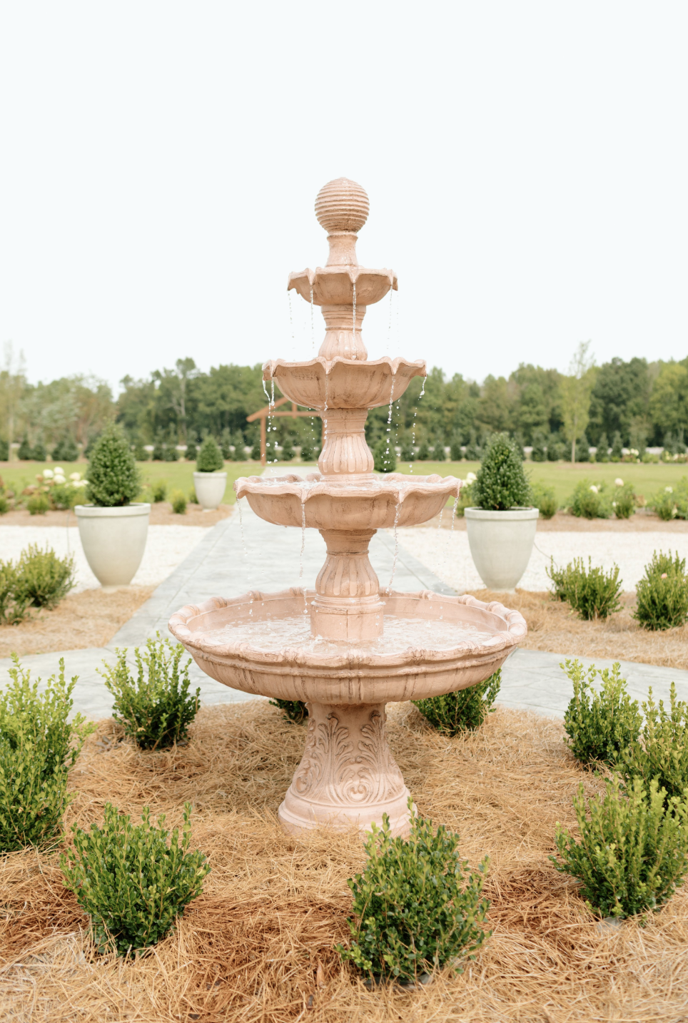 Pink stone tiered fountain in landscaped garden with potted plants and trees in the background.