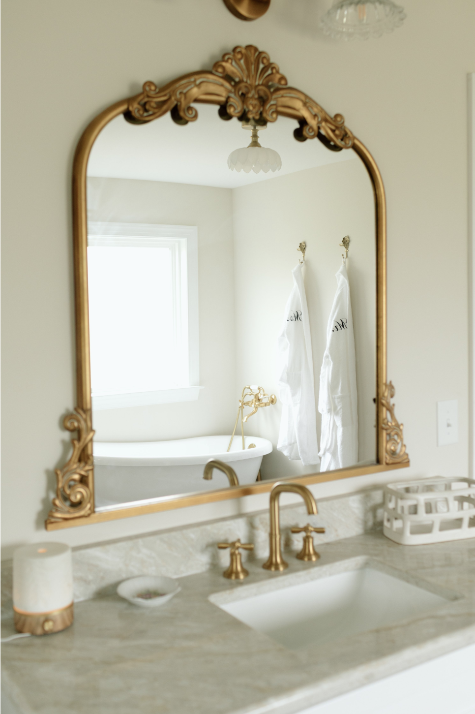 Bathroom with a marble sink, gold fixtures, an ornate gold mirror, a window, a white bath with gold fixtures, two white robes hanging on hooks, and a white basket on the counter.