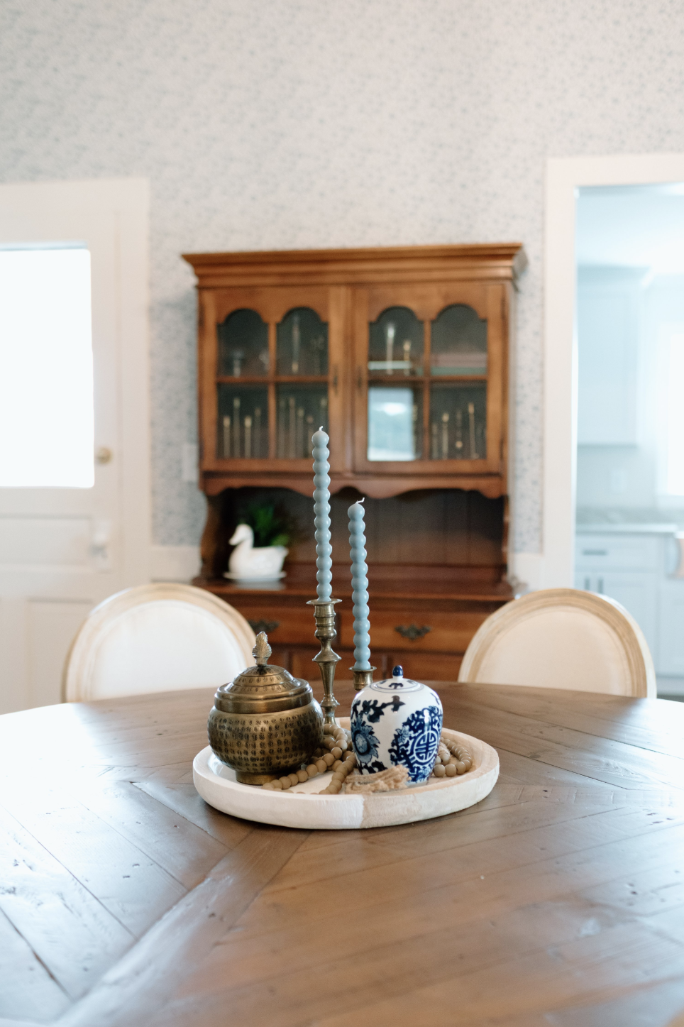 Decorative centerpiece with candles, a jar, and beads on a wooden dining table in a room with a wooden cabinet and patterned wallpaper.