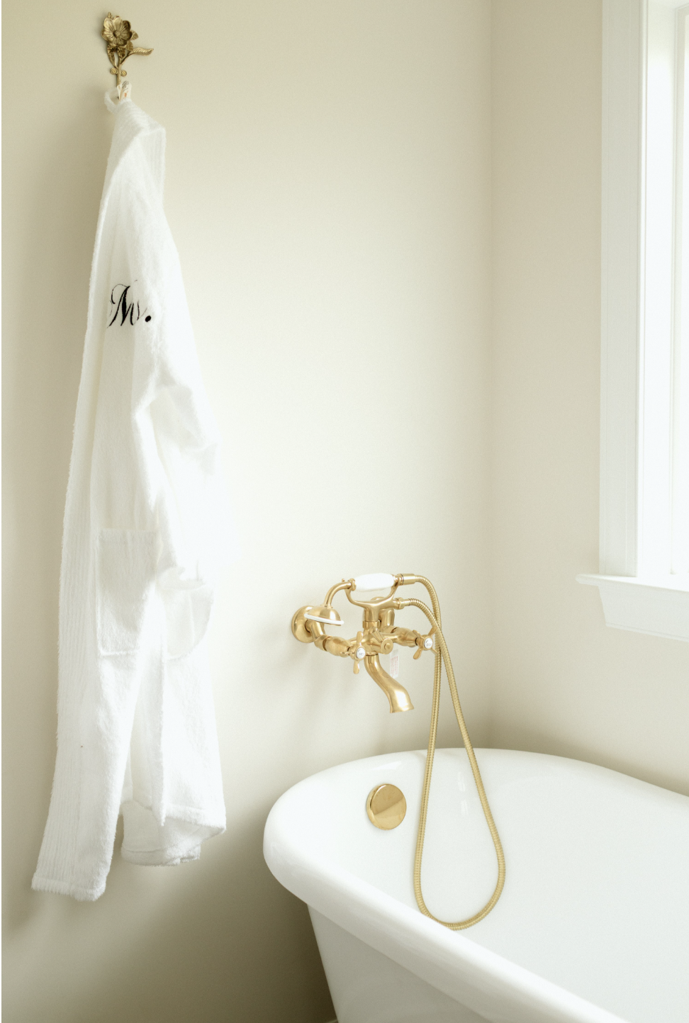 A white towel hanging on a decorative gold hook on a pale yellow wall near a bathtub with gold fixtures and a window with white trim.