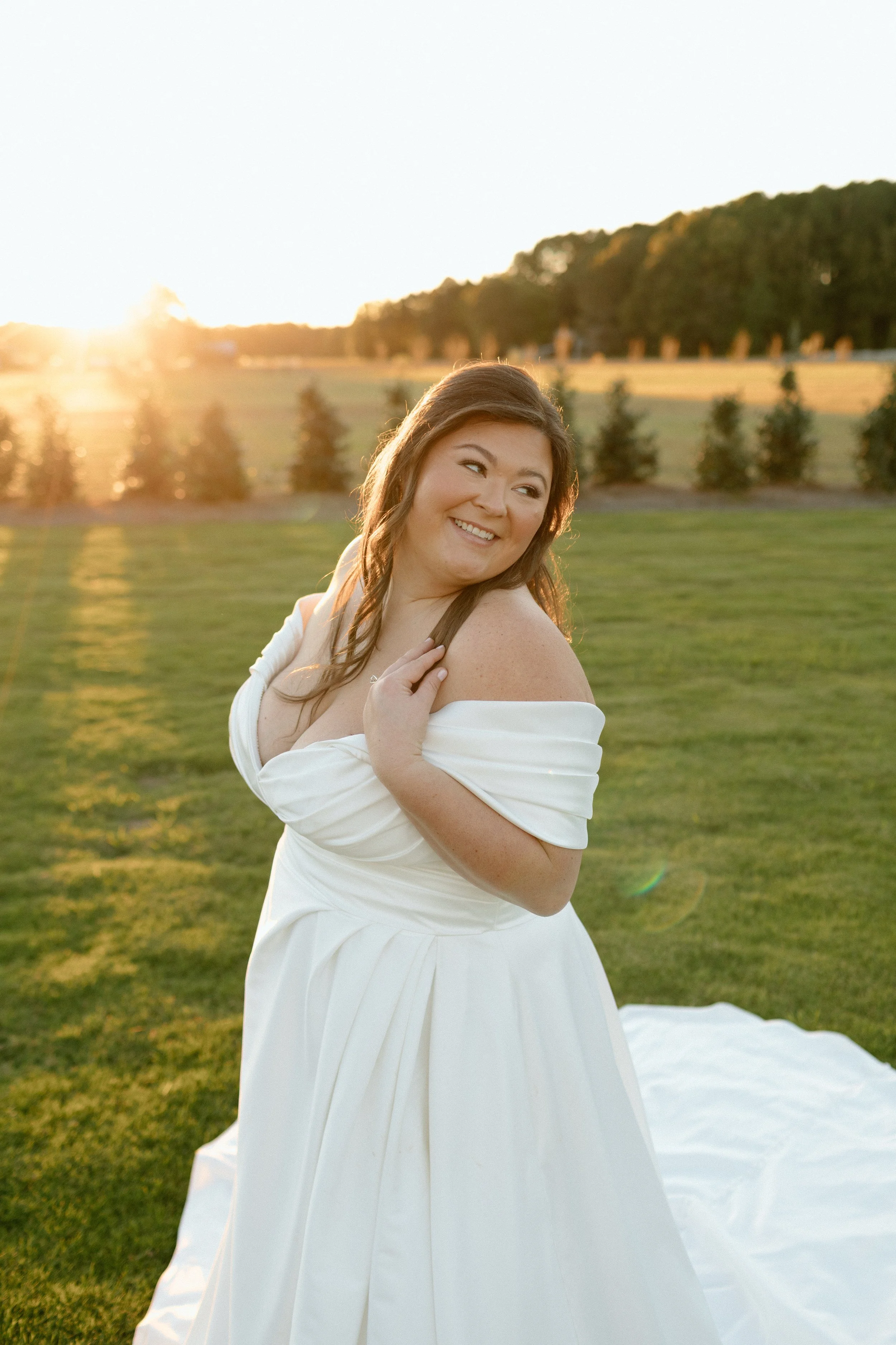 A woman in a white off-shoulder wedding dress smiling outdoors during sunset with a grassy field, trees, and a row of wind turbines in the background.