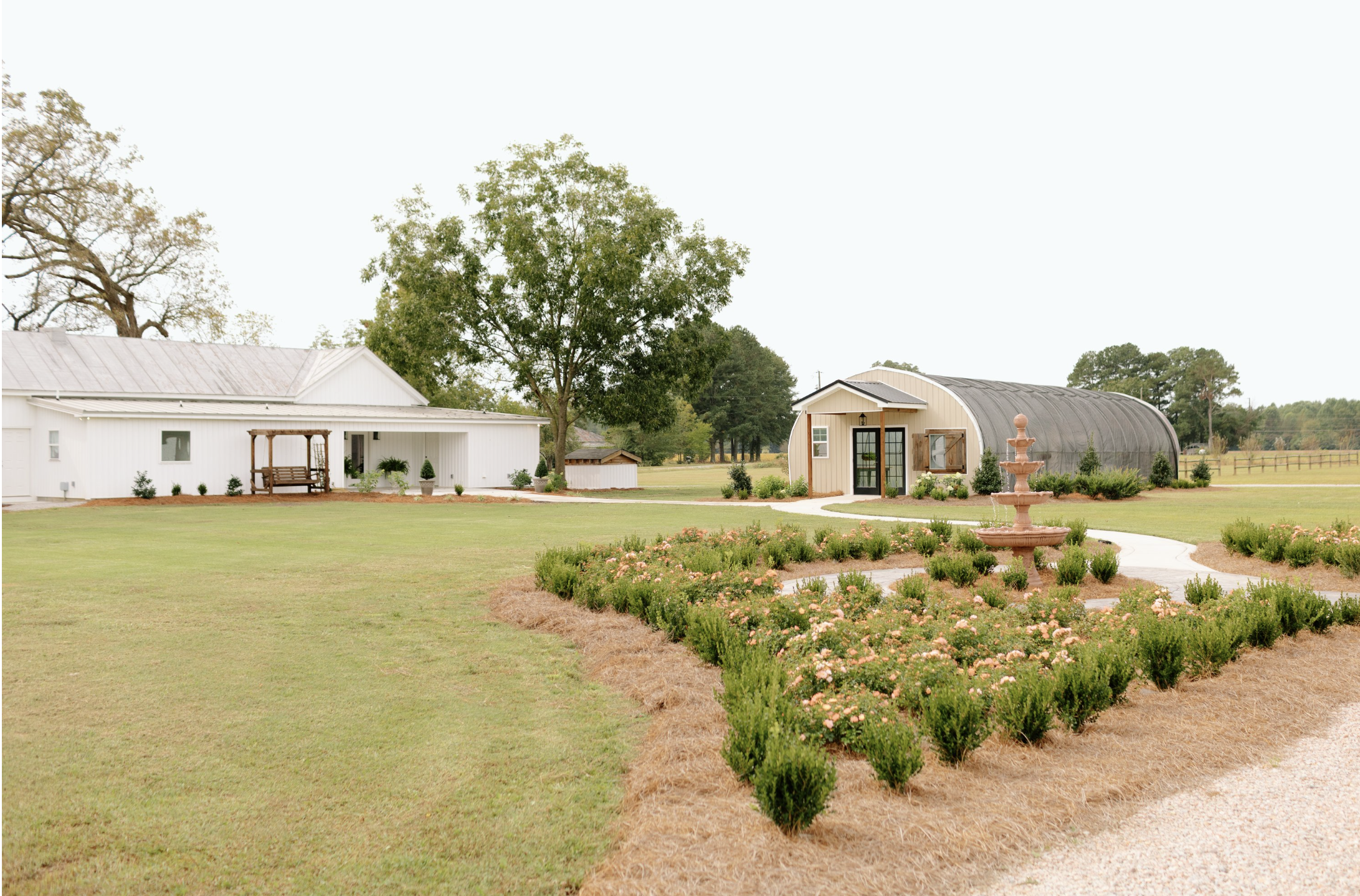 A rural landscape featuring a manicured lawn, a flower garden with a tiered fountain, two buildings (a white one with a porch and a beige one with a curved roof), and trees in the background under a cloudy sky.