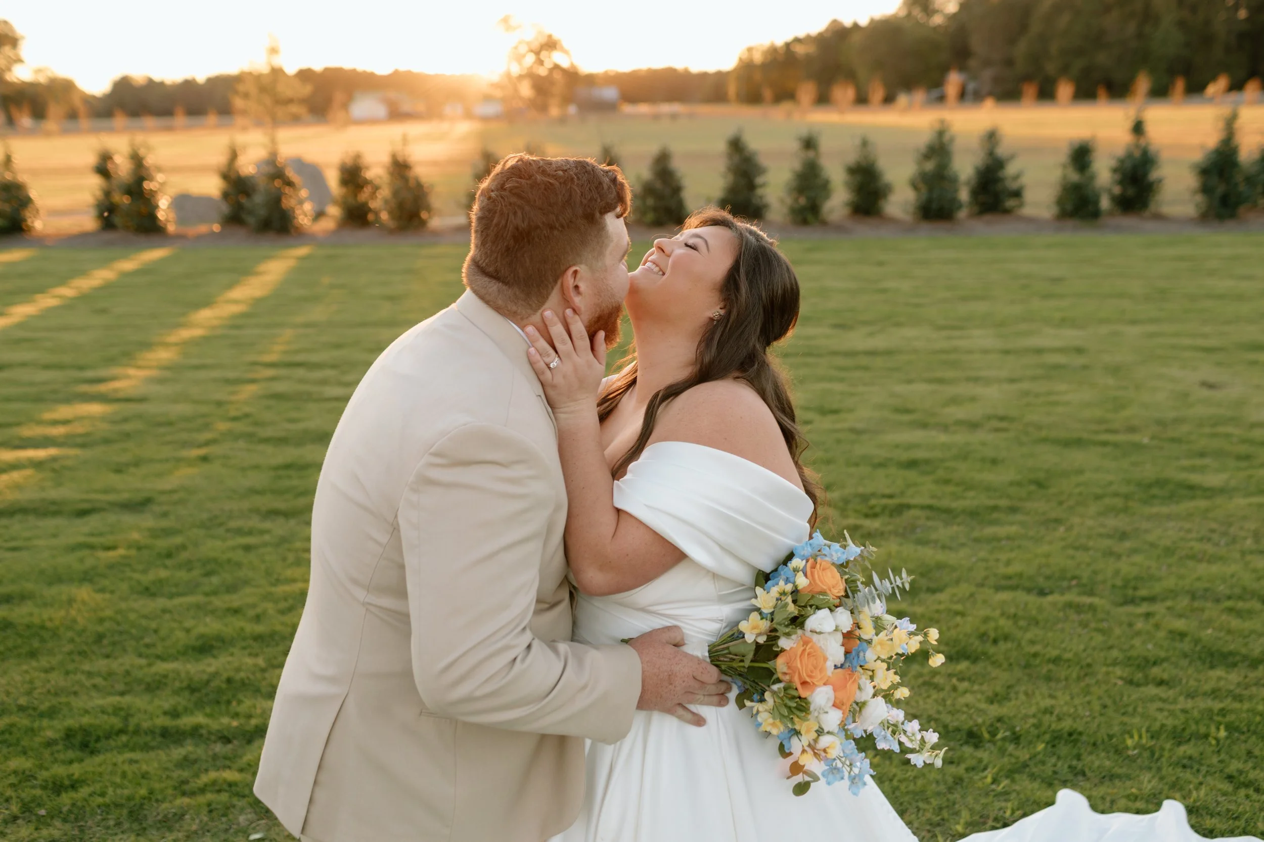 A newlywed couple embracing and smiling outdoors at sunset, with a green lawn, blooming flowers, and trees in the background.