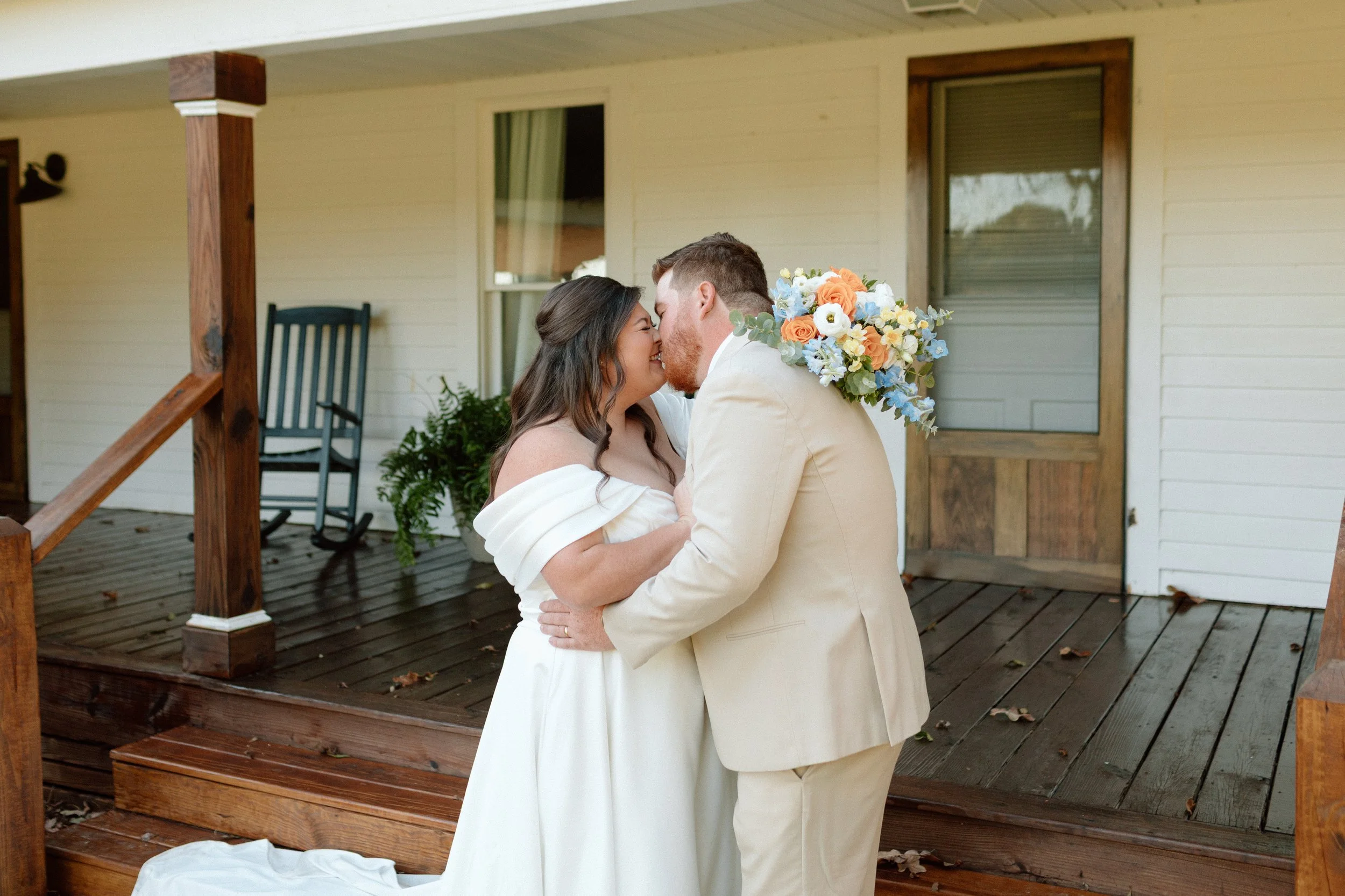 A couple dressed in wedding attire, smiling and leaning in for a kiss on a wooden porch outside a house with yellow siding.