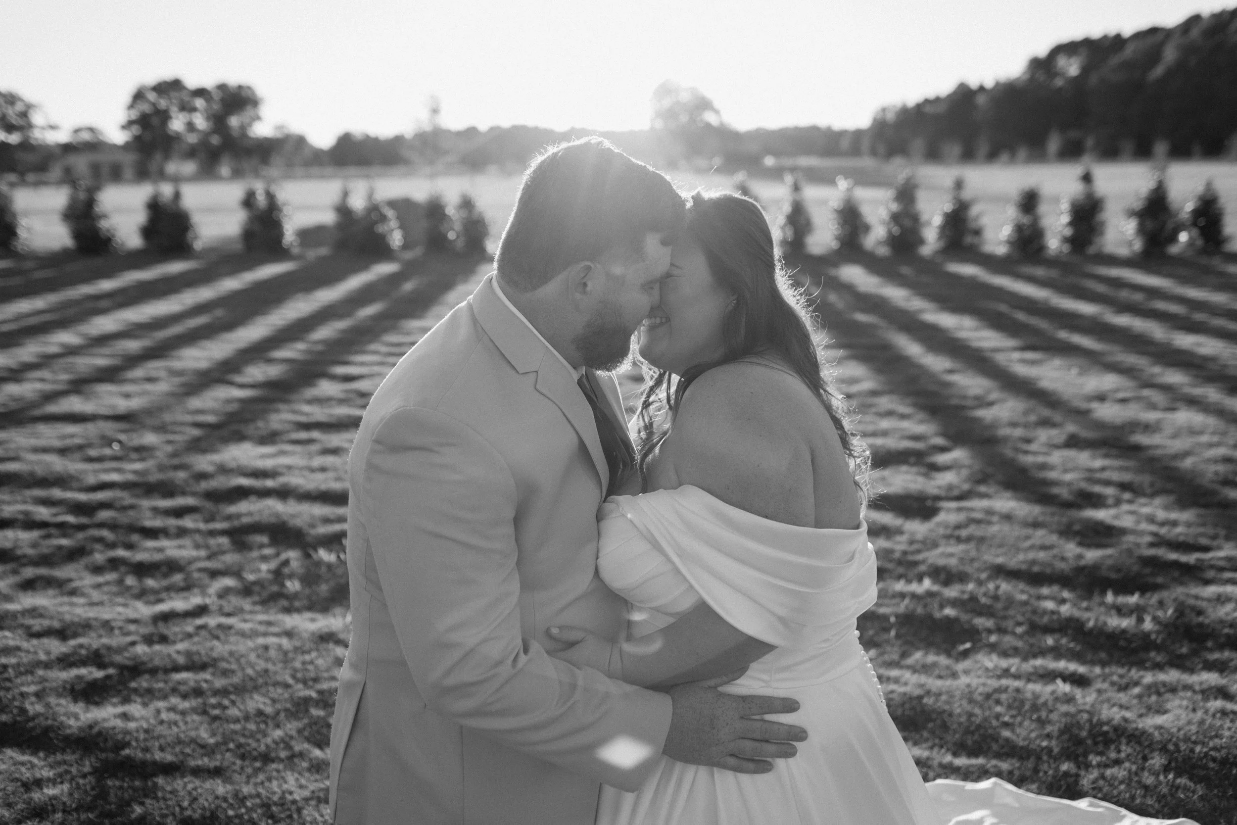 A black-and-white photo of a couple dressed in formal attire embracing and about to kiss outside in a field with trees and a fence in the background, with sunlight creating a backlit effect.