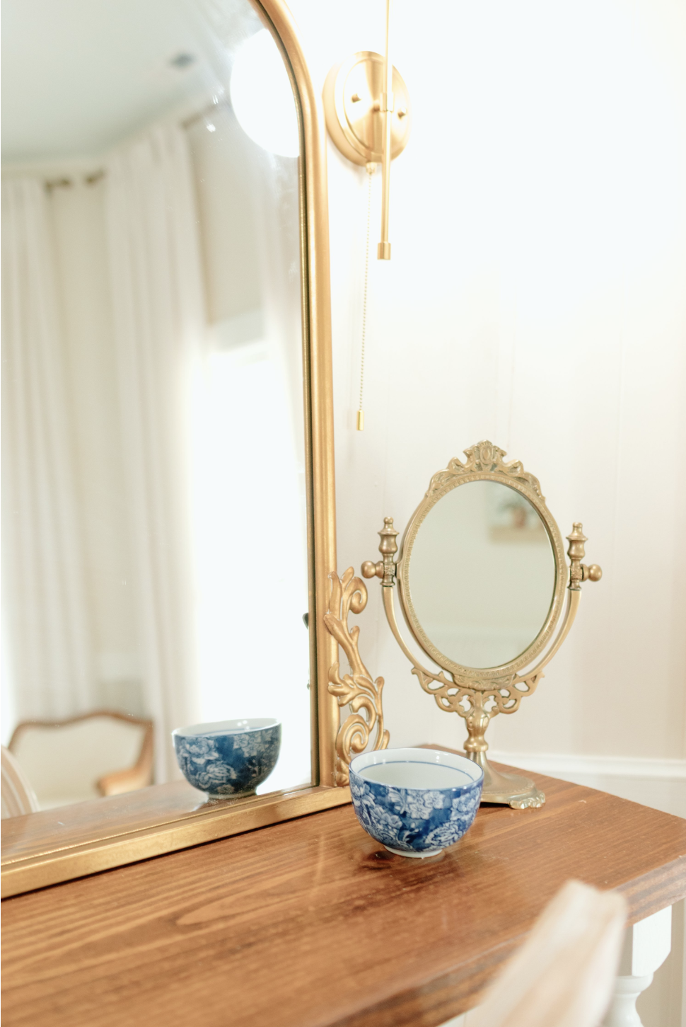 A wooden dresser with a gold-framed mirror attached, a small ornate gold-framed vanity mirror, and two blue and white porcelain bowls. A wall sconce with a pull chain is mounted above, and a glimpse of a chair with a wooden frame and yellow upholster