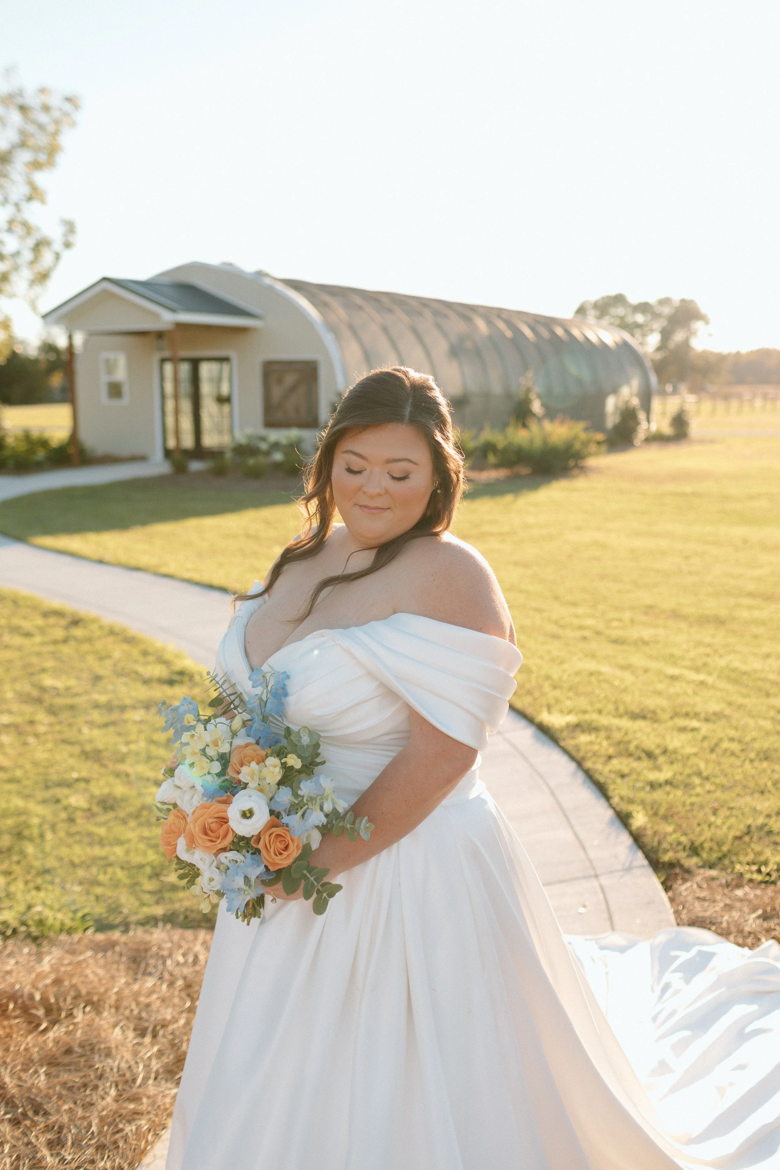 A woman in a white wedding dress holding a bouquet of flowers outdoors during sunset, with a building and trees in the background.