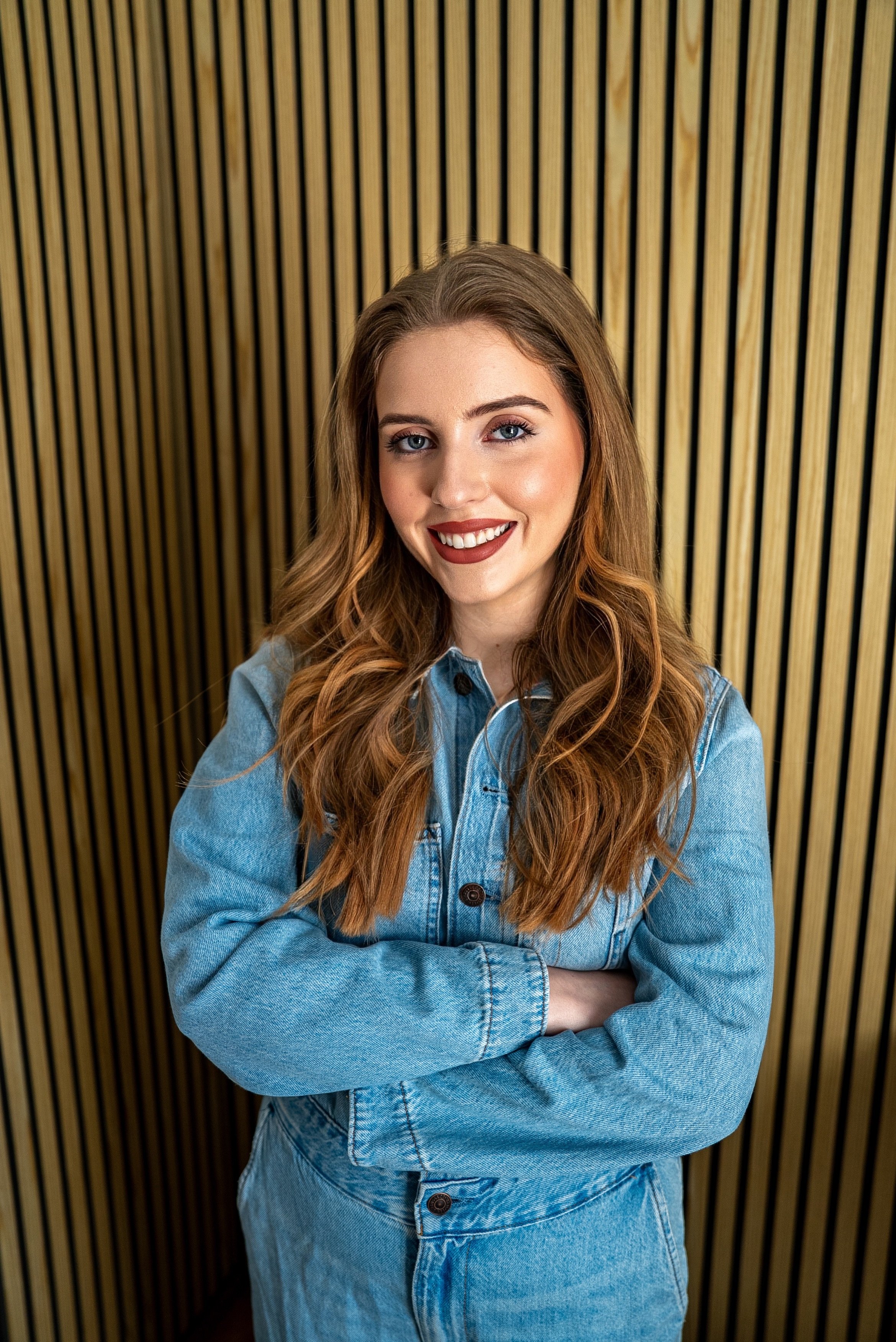 A young woman with long red hair, blue eyes, and light makeup, standing with her arms crossed in front of a wood-paneled wall, smiling and wearing a light blue denim jacket.