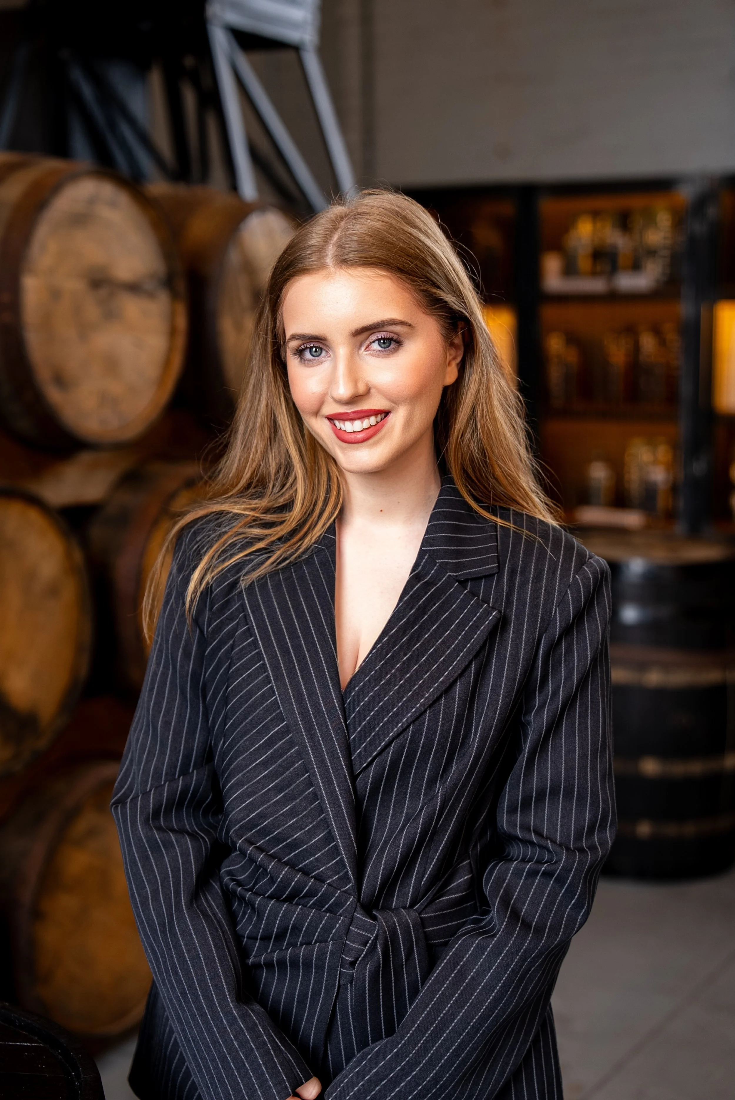 A young woman with long, wavy blonde hair and blue eyes, smiling and wearing a black pinstripe blazer, standing in front of stacked wooden barrels in a dimly lit cellar or bar setting.