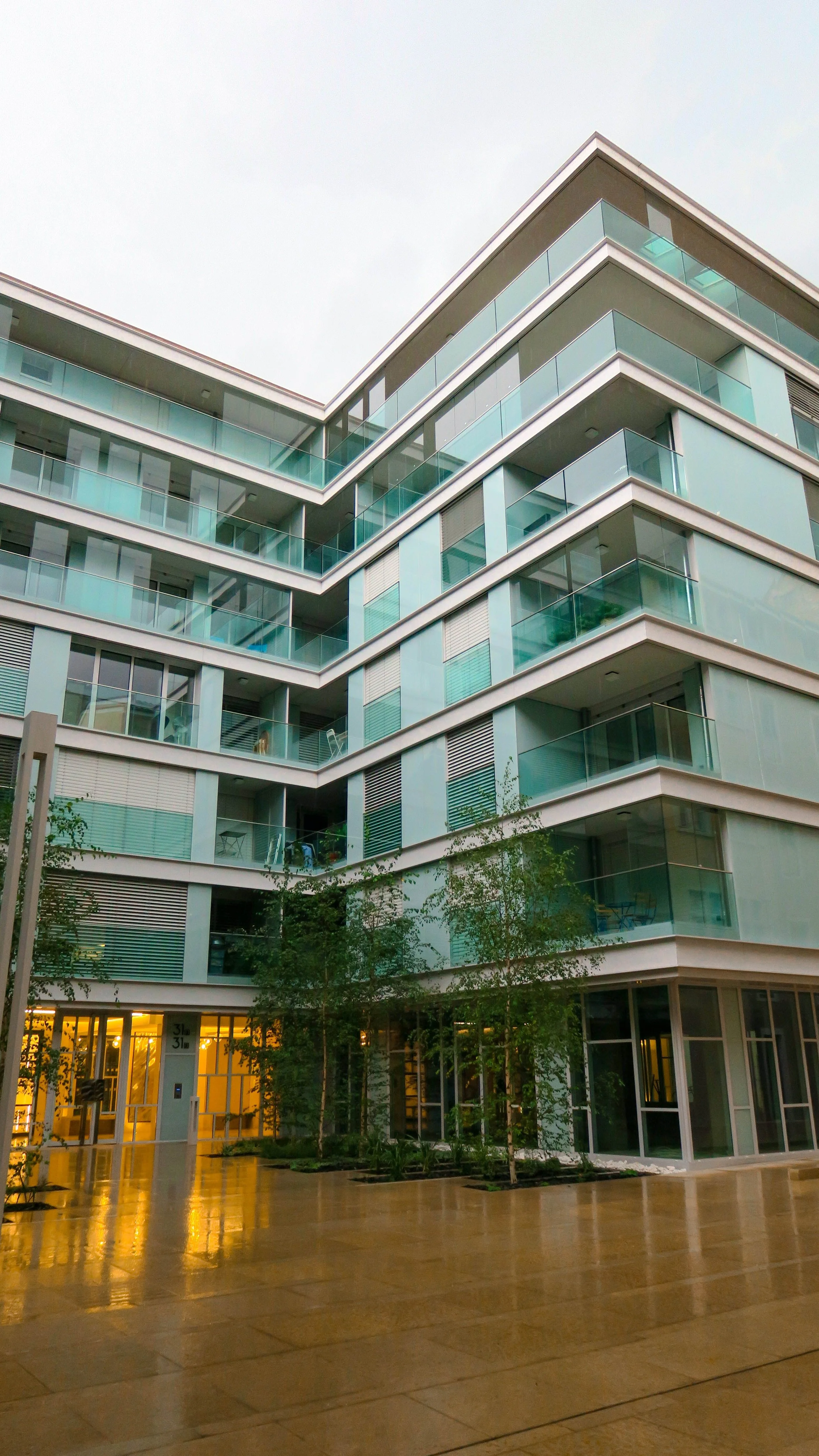 Modern multi-story residential building with glass balconies and large windows, surrounded by trees and a reflective tiled courtyard.