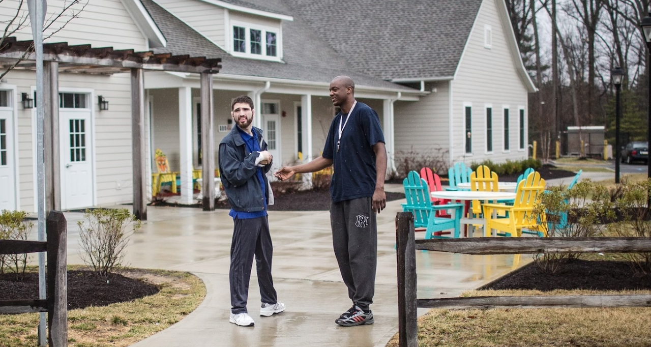 Two men standing outside near a building, shaking hands and smiling. One man is wearing a jacket and the other is in a T-shirt. The setting appears to be a residential community with colorful outdoor chairs and tables in the background, on a wet day.