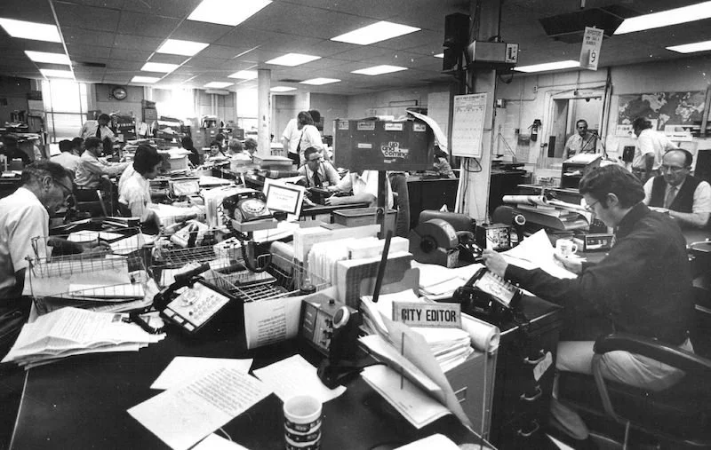 Black and white photo of a busy newsroom with numerous desks filled with papers, computers, typewriters, and telephones, with reporters and editors working diligently.
