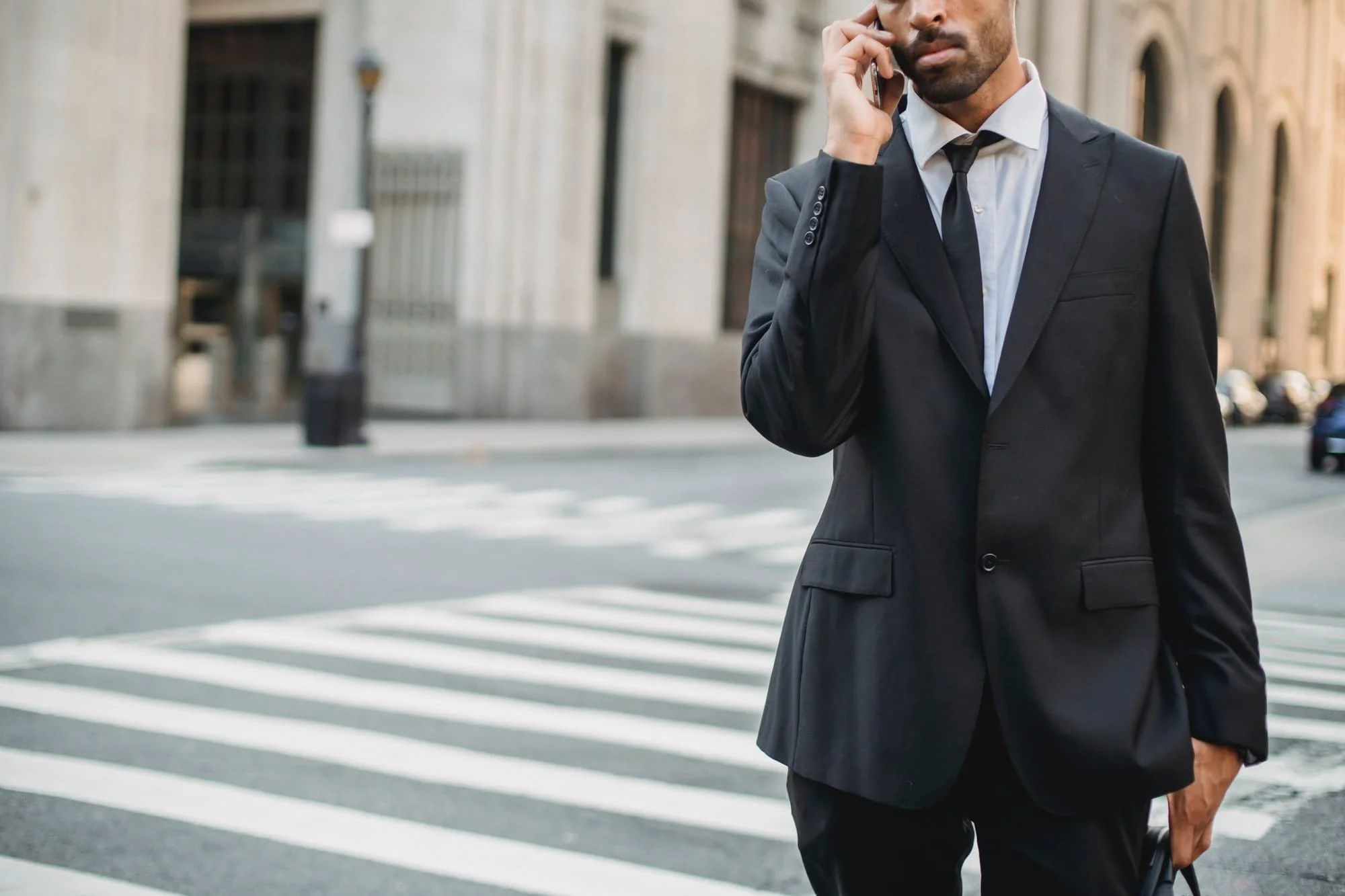 A man in a black suit with a white shirt and black tie standing on a city street, talking on his cellphone.