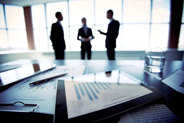Three business professionals having a discussion in a modern office with large windows, while documents and a tablet displaying a bar graph are on the table in the foreground.