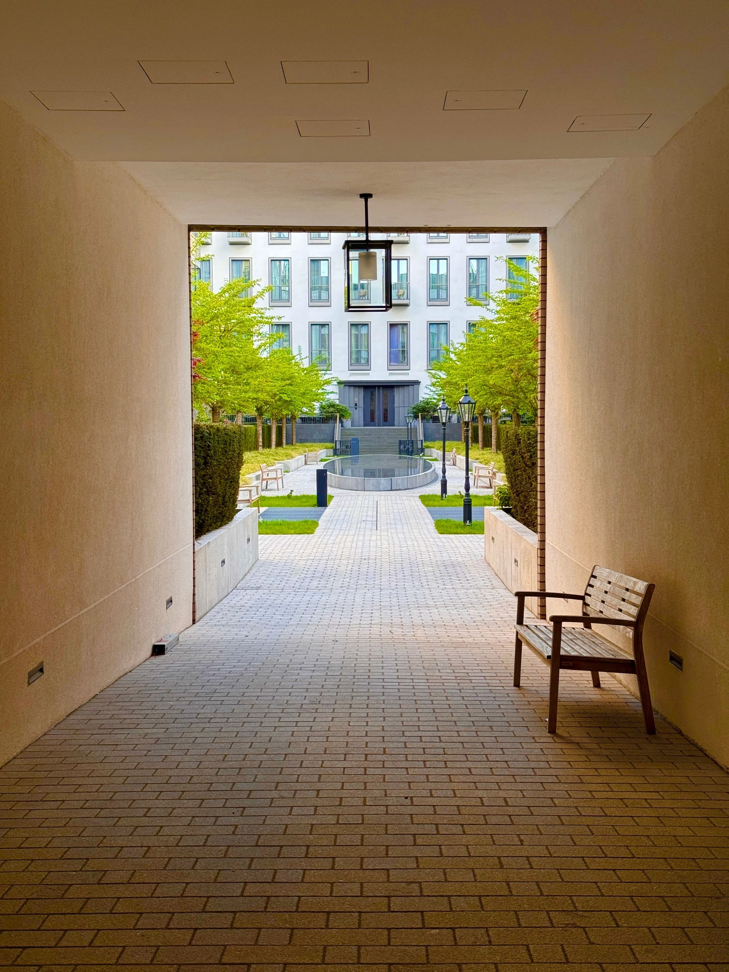 View from a covered walkway opening onto a courtyard with benches, trees, lamp posts, and a modern building in the background.