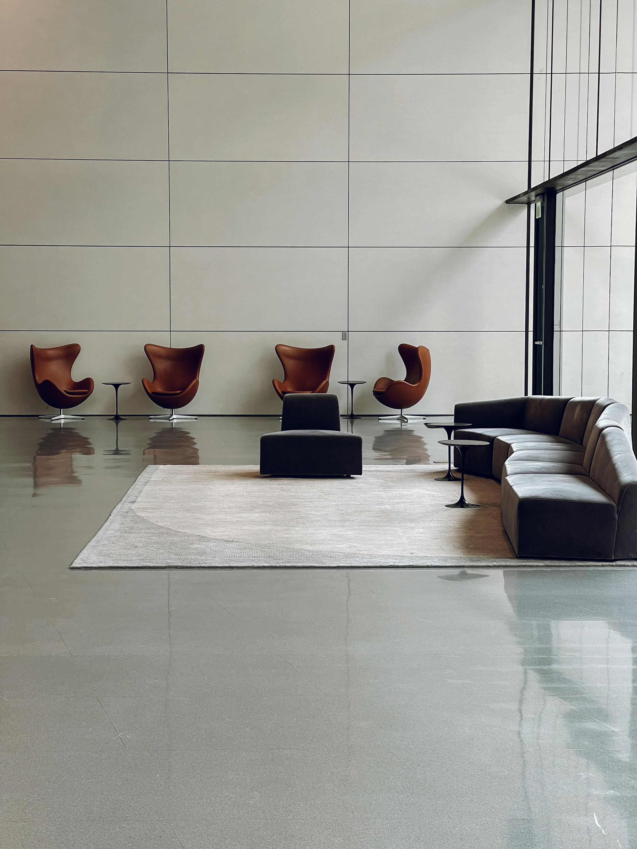 Modern indoor waiting area with four brown chairs, one black ottoman, a curved gray sofa, and small black side tables, with a large window and tiled wall.