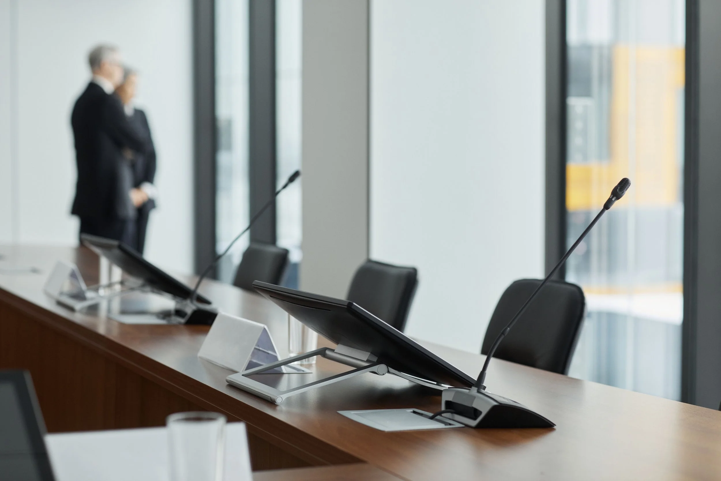 Empty conference room with black chairs, microphones, and nameplates on a long table. Large windows in the background.