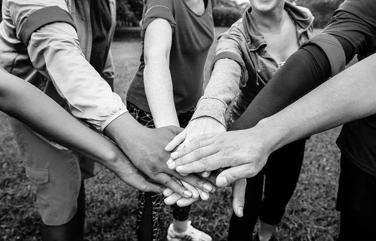 Group of diverse people with hands stacked together in the center, outdoors, symbolizing unity and teamwork.