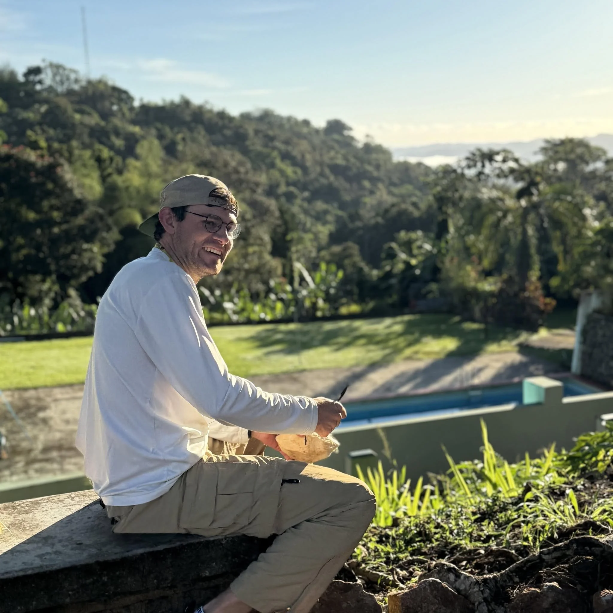 A man sitting on a stone bench outdoors, smiling and wearing a beige cap, glasses, a white long-sleeve shirt, and khaki pants, with a background of trees and grass during daylight.