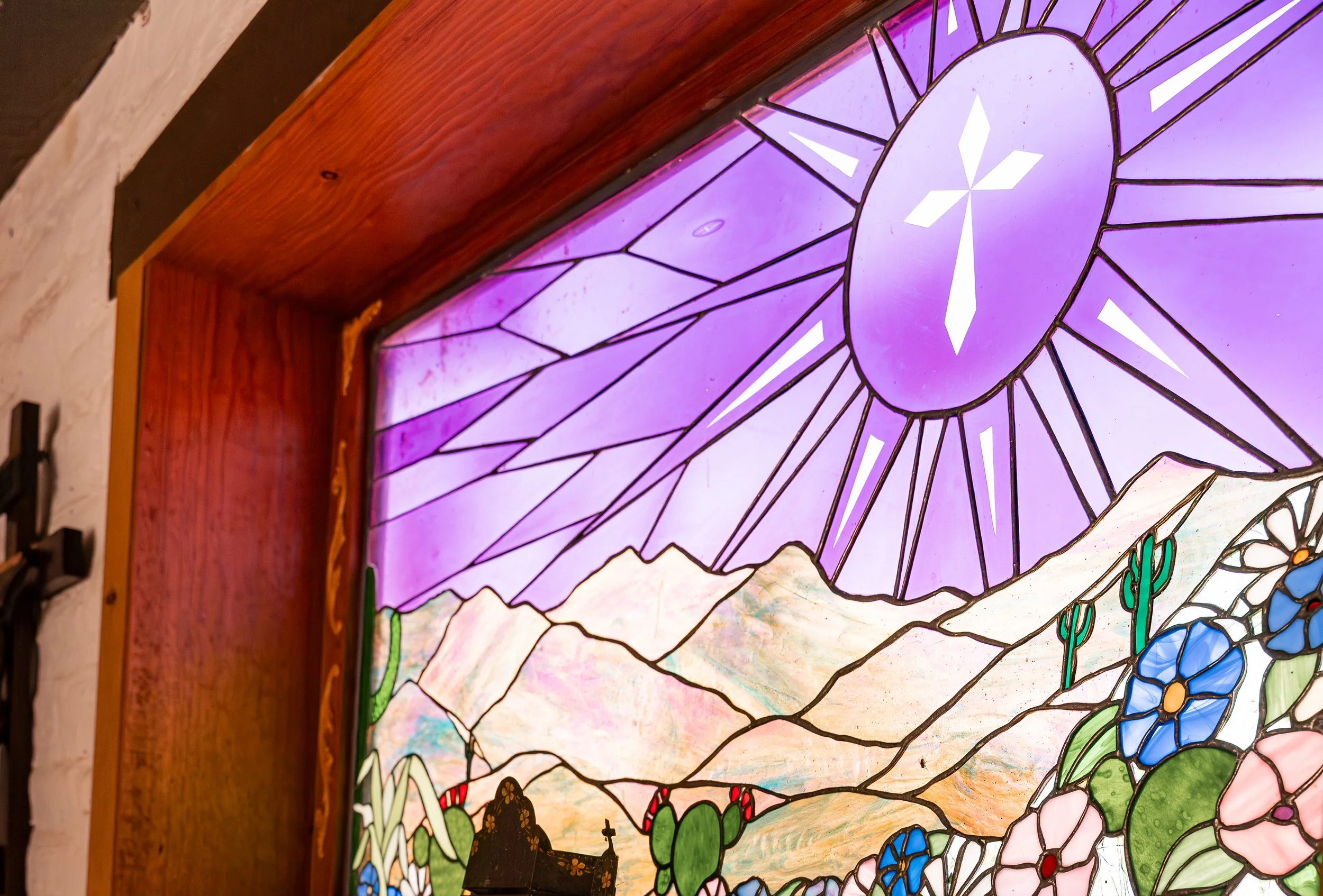 A stained glass window with a mountain scene, featuring a prominent purple mountain and the sun shining behind it, framed in wood.