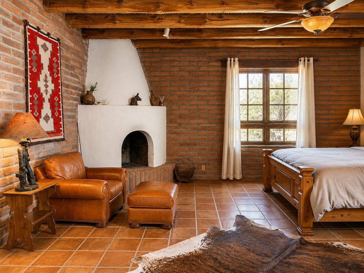 A rustic bedroom with exposed red brick walls, a large window with white curtains, a wooden bed with a beige blanket, a leather armchair with an ottoman, a cowhide rug, and a white stucco fireplace with decorative items on the mantel.