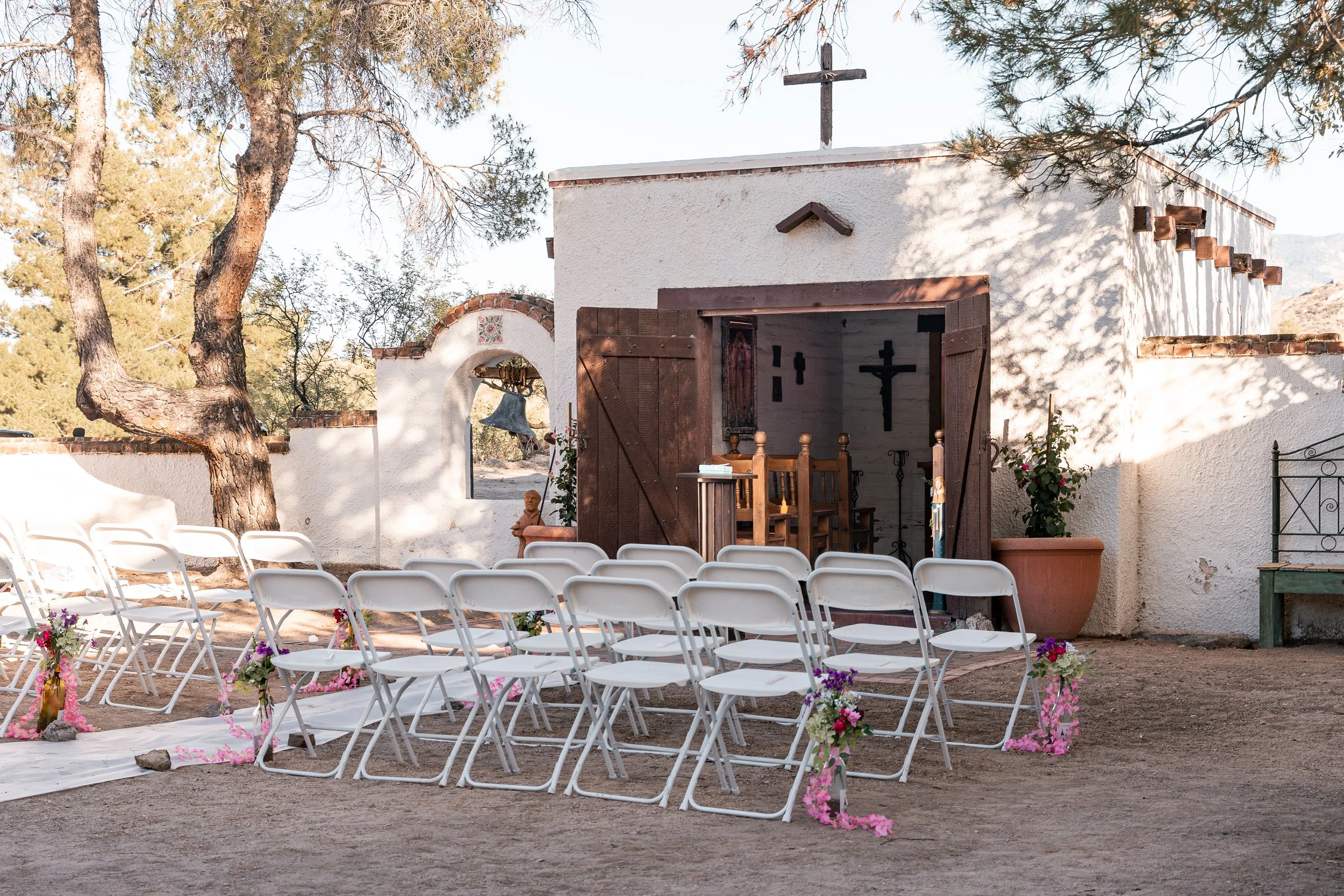 Outdoor small church with white stucco walls, wooden doors, and a cross and a small roof on top. The church is decorated for a wedding with white chairs arranged in rows, pink and purple flowers, and a white aisle runner. There is a large tree to the left, and a bell hanging near a small archway on the left side of the church entrance.