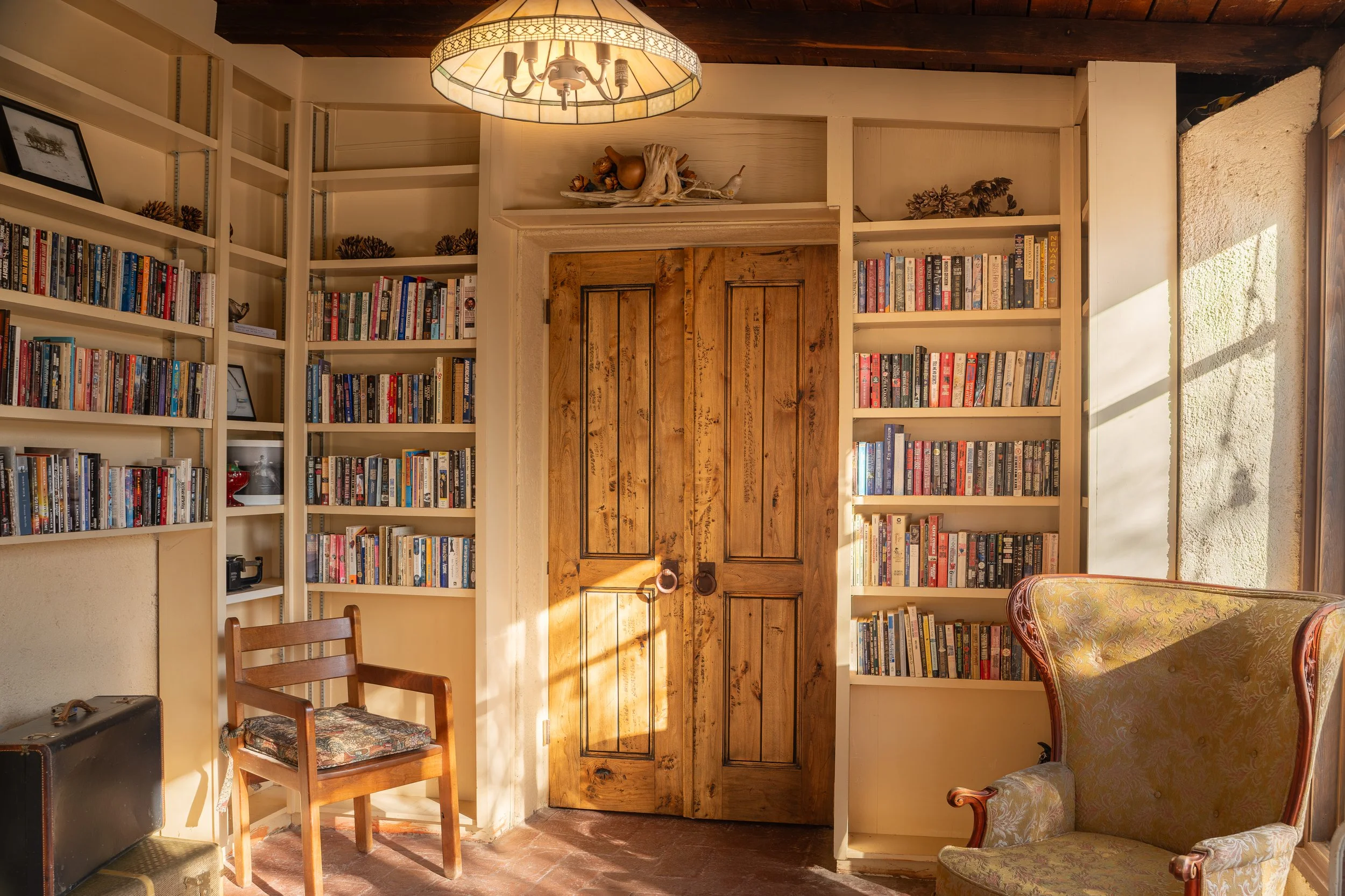 A cozy living room with built-in white bookshelves filled with books, a wooden door, a chandelier hanging from the ceiling, a wooden chair, an upholstered armchair, and decorative items like pinecones and wooden ornaments.