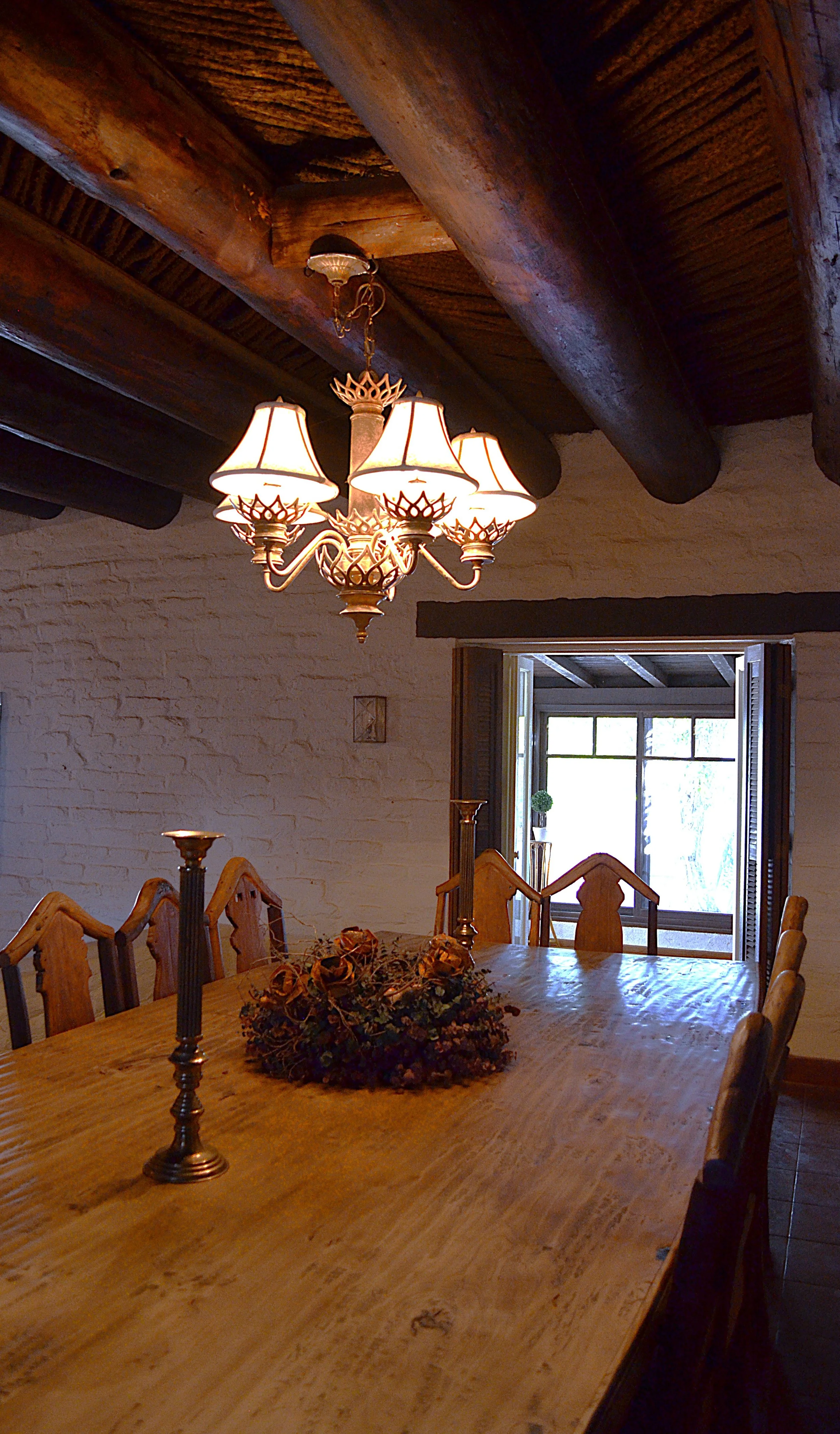 Interior of a rustic dining room with a wooden table, a chandelier, and a window showing outdoor scenery.