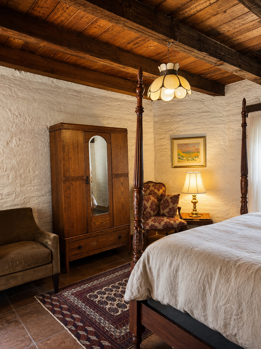 A cozy bedroom with a four-poster bed, vintage wooden wardrobe, armchair, patterned rug, side table with a table lamp, framed artwork on a white brick wall, and a window with curtains, wooden ceiling beams.