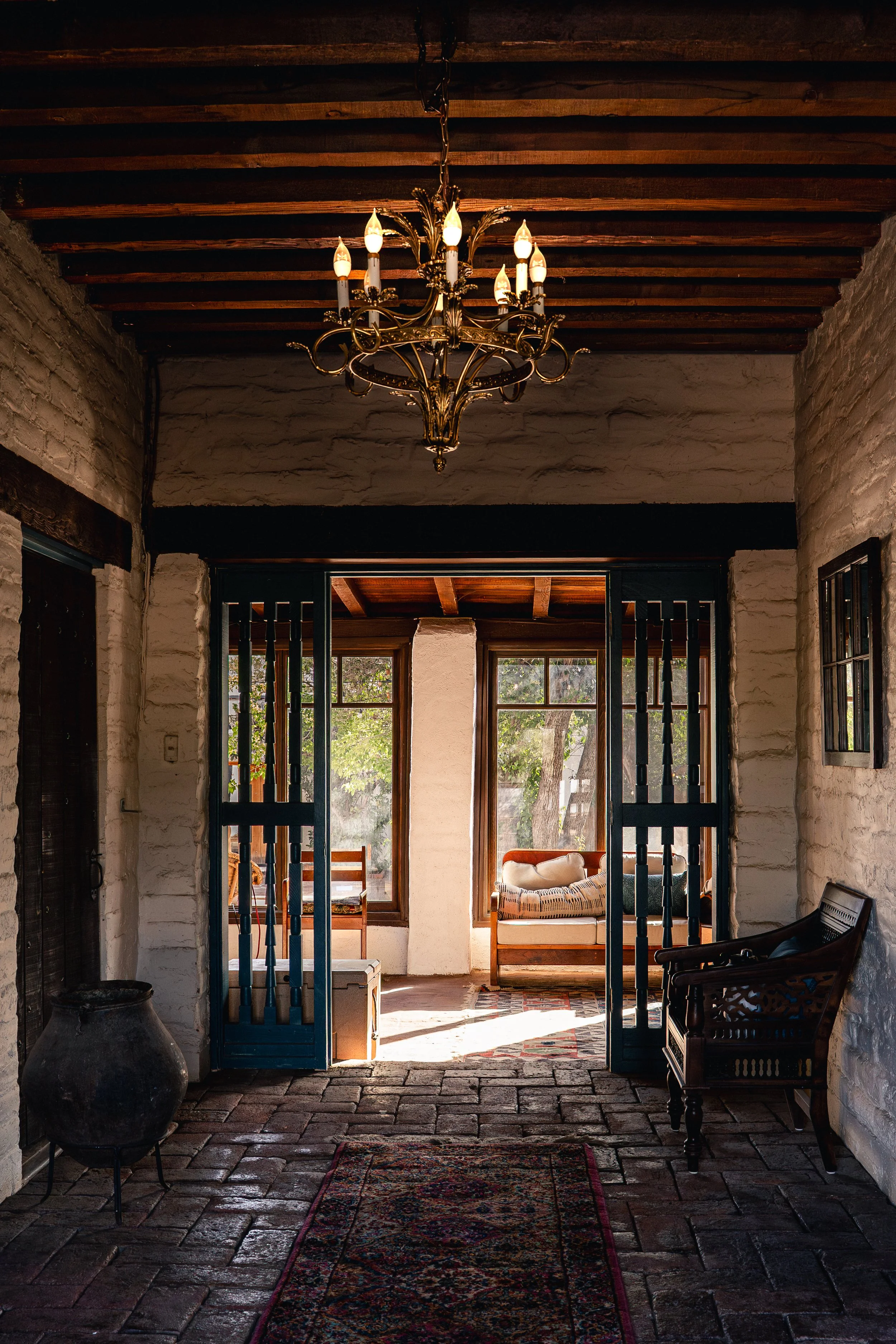 A cozy interior view of a rustic room with a chandelier hanging from a wooden ceiling, brick walls, and a doorway leading to a sunlit porch with chairs and a sofa.