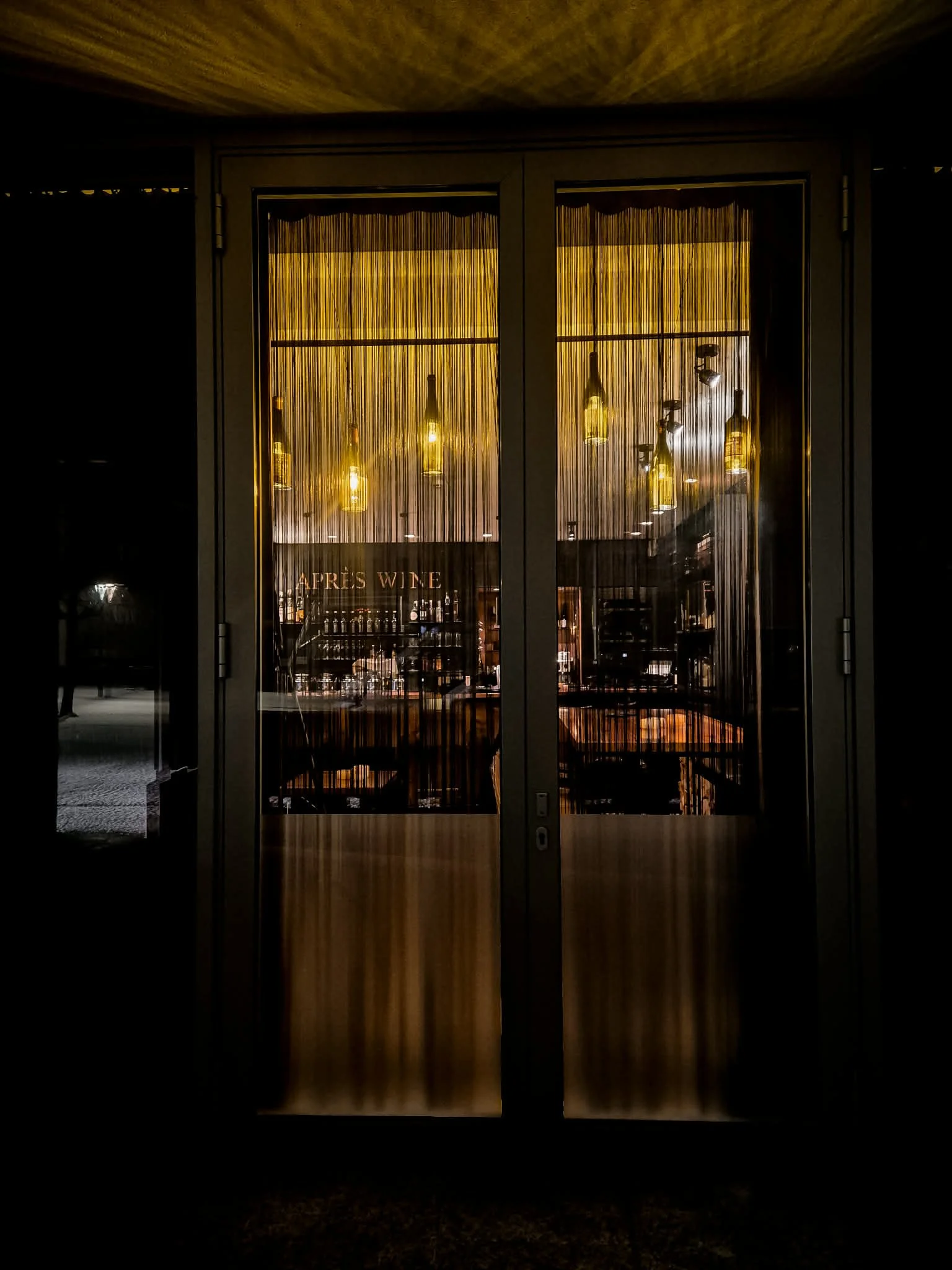 A view through glass doors featuring string curtains and hanging lights, with a sign that reads 'APRE'S WINE' visible inside.