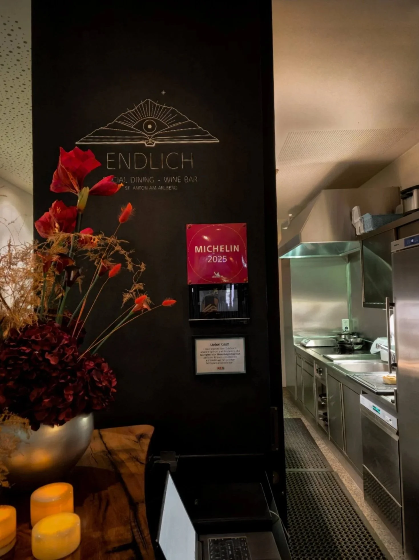 Interior of a restaurant or café, showing a black wall with a logo that includes an open book, an eye, and stars, and the word 'ENDLICH' underneath. There is a decorative floral arrangement with red flowers in the foreground and a sign mentioning 'Mi