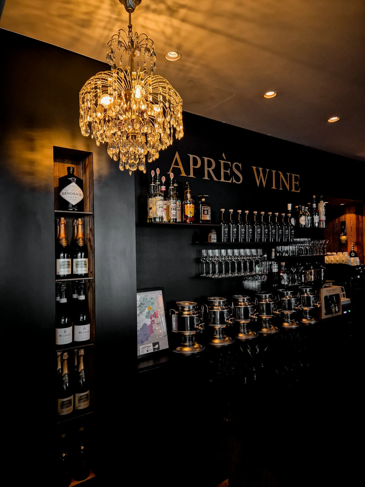 Interior of a bar with a chandelier hanging from the ceiling, bottles of alcohol on shelves, and a sign that reads 'APRÈS WINE' on the wall.