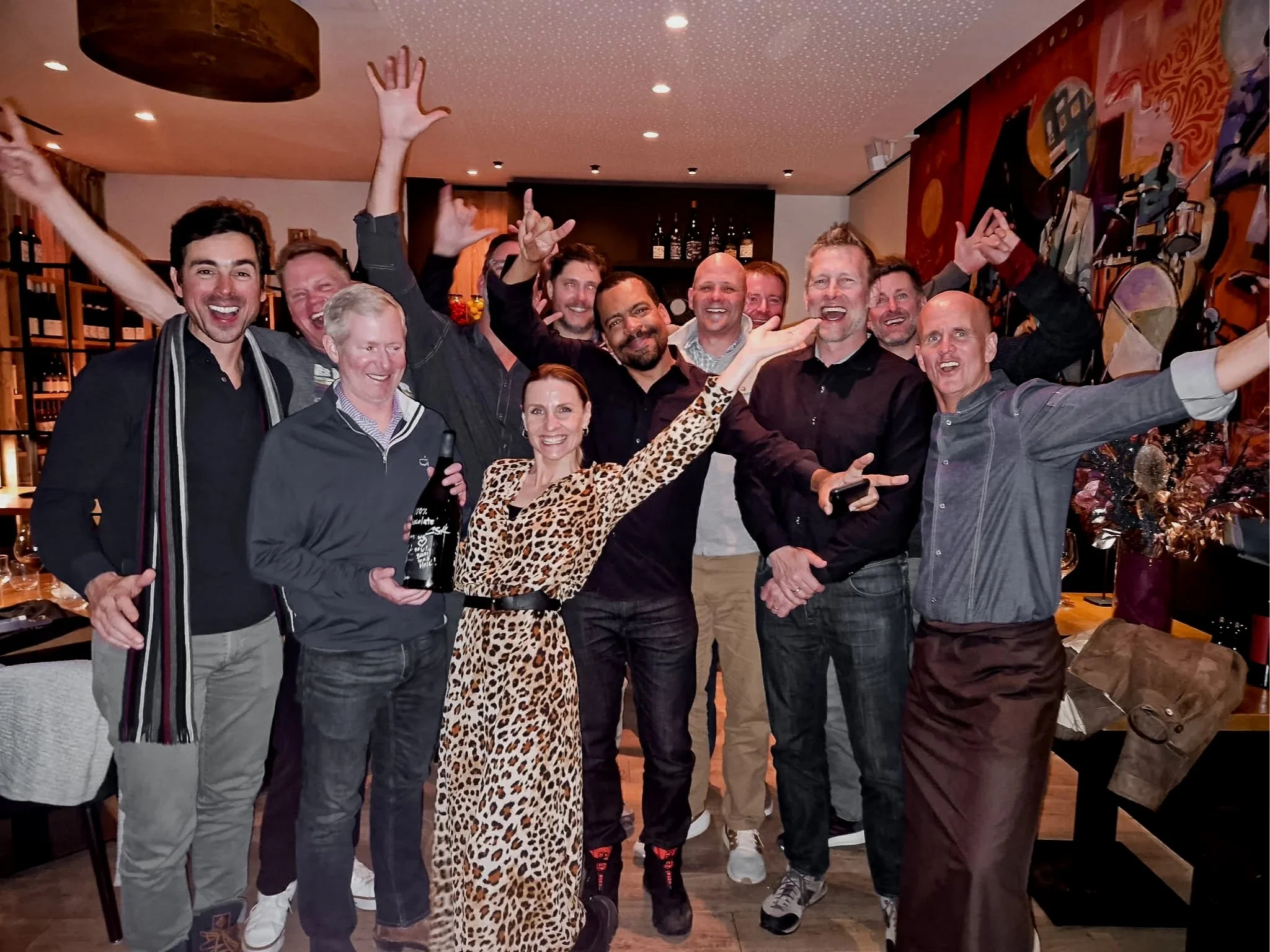 Group of people celebrating and smiling, posing together with some holding drinks, in a cozy indoor setting with wine bottles on shelves and colorful artwork on the wall.