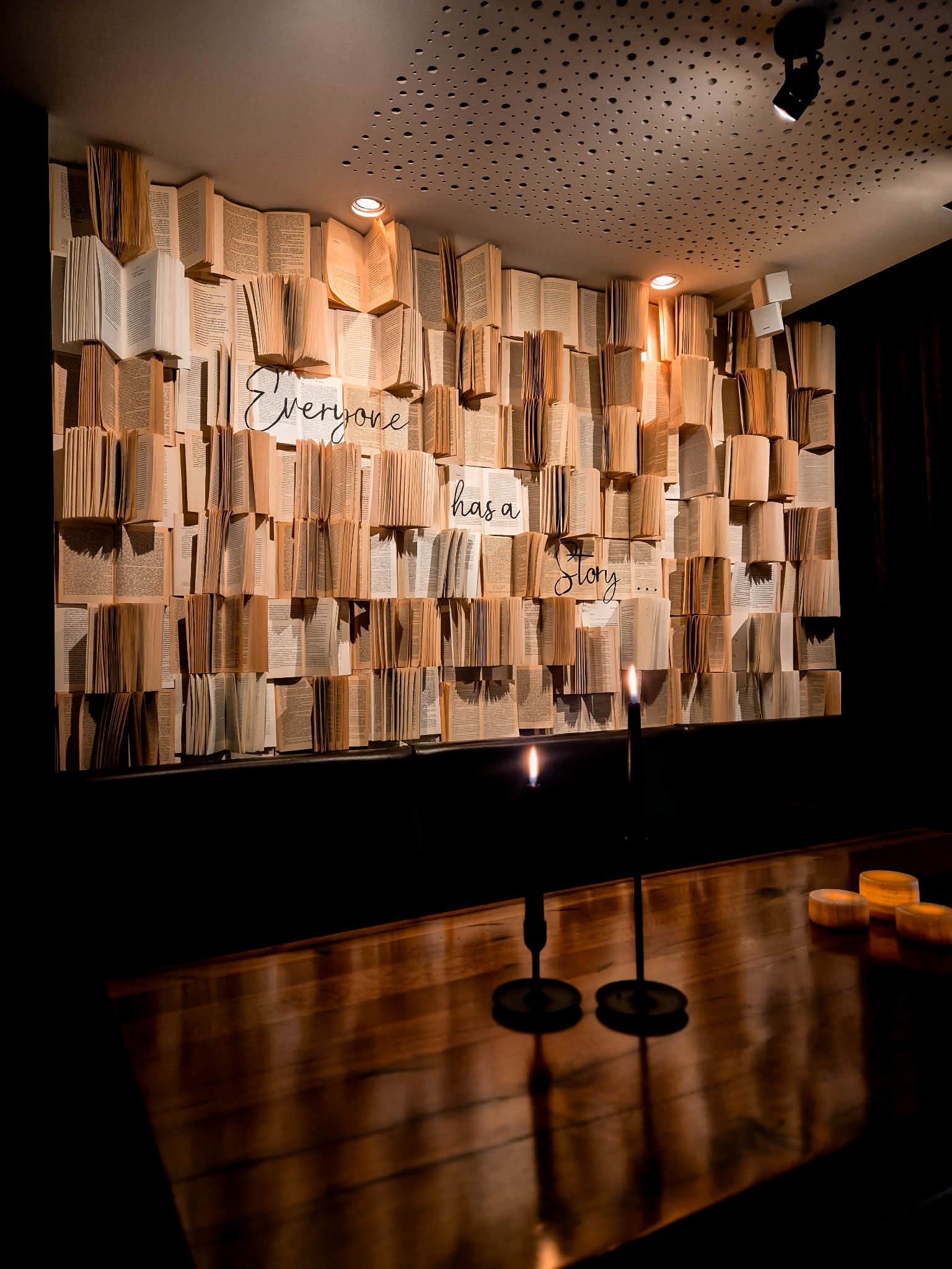 Display of open books on a wall with the words 'Everyone has a story' written among them, situated in a dimly lit space with candles and spotlights.