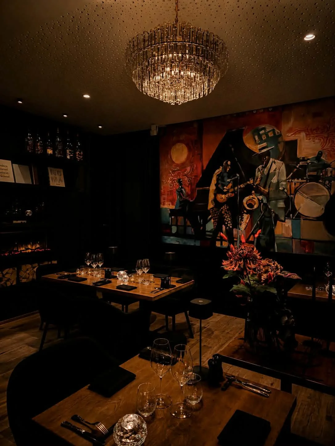 A dimly lit upscale restaurant with a modern chandelier, a colorful mural of jazz musicians, a dining table set with wine glasses, plates, silverware, and candles, and a large floral arrangement.