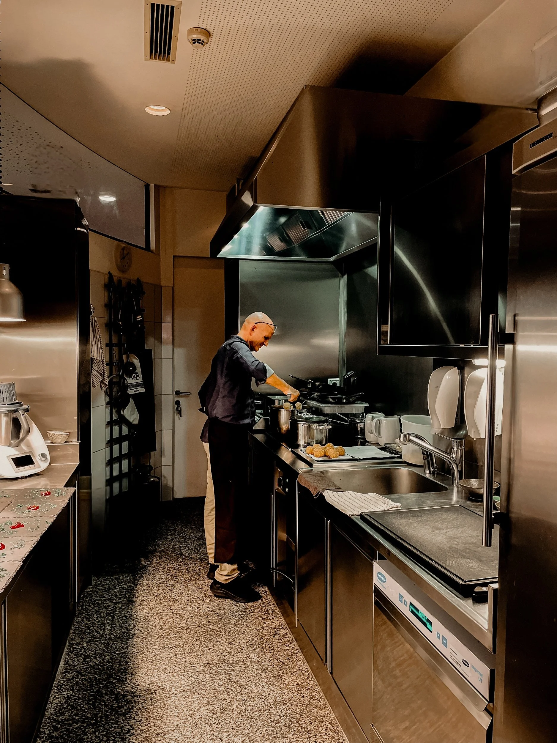 A man cooking in a commercial kitchen, standing at a stove with pots and frying food, with kitchen appliances and utensils around him.