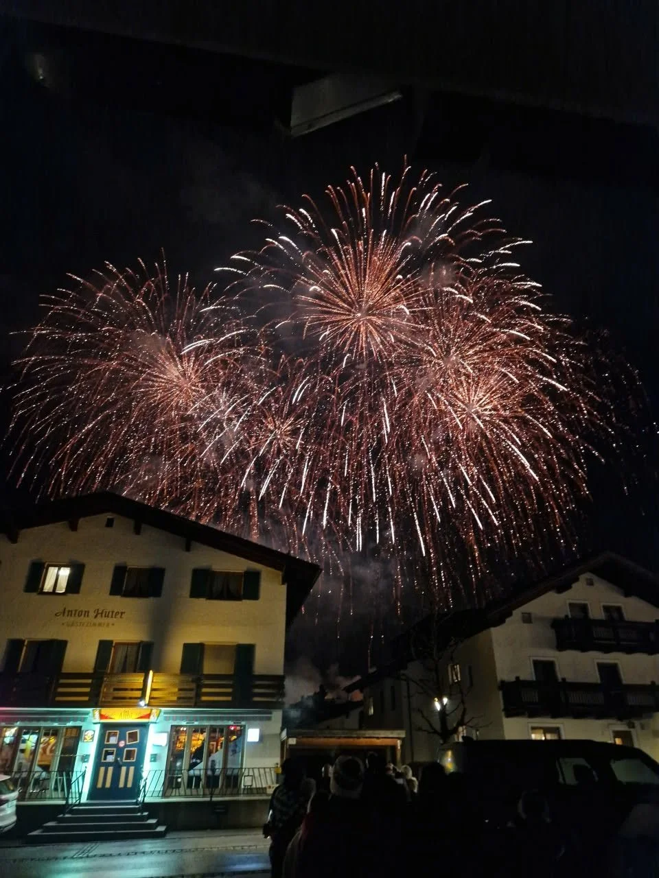 Night sky with large fireworks exploding above buildings and people watching.