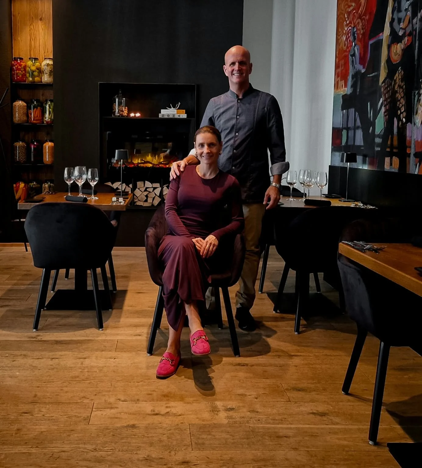 A man and woman in a restaurant with empty wine glasses on the tables. The woman is sitting in a chair, wearing a maroon dress and pink shoes, while the man stands behind her with his hand on her shoulder, smiling. There is a fireplace, art, and shelves with jars in the background.