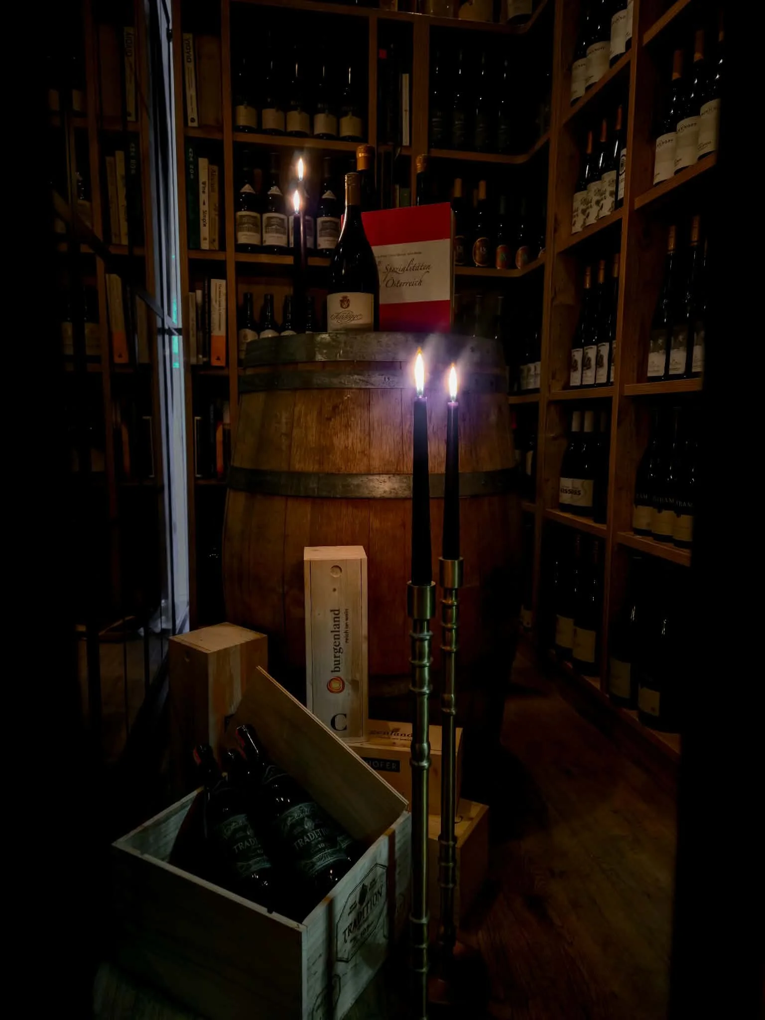 Dimly lit wine cellar with wooden shelves filled with wine bottles. In the center, a wine barrel displays a bottle of wine and a red menu or sign. Two lit candles stand in front of the barrel, casting a warm glow over the scene.