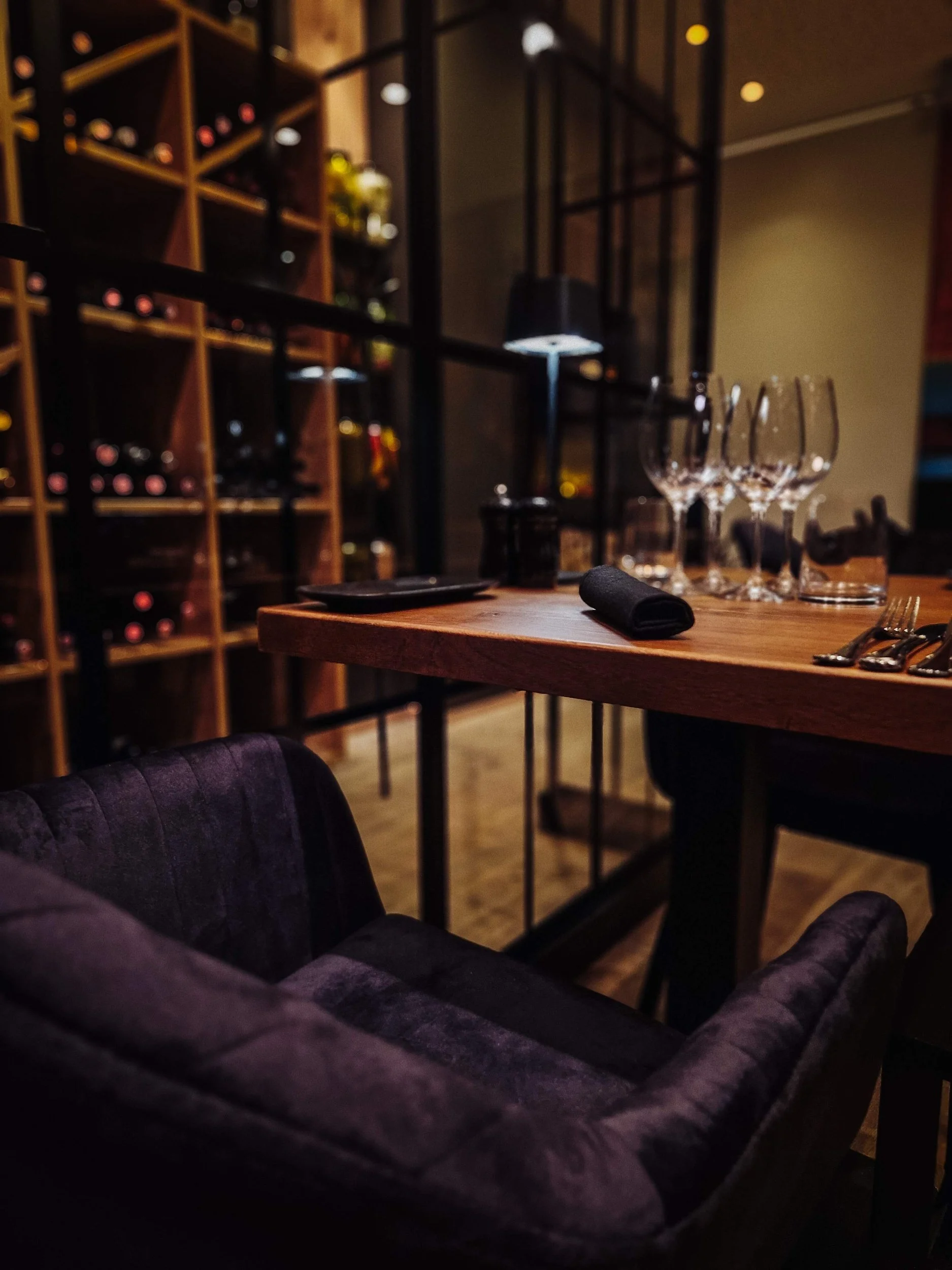 A dimly lit restaurant table set with four empty wine glasses, a black cloth napkin, and silverware, with a wine rack filled with bottles in the background.