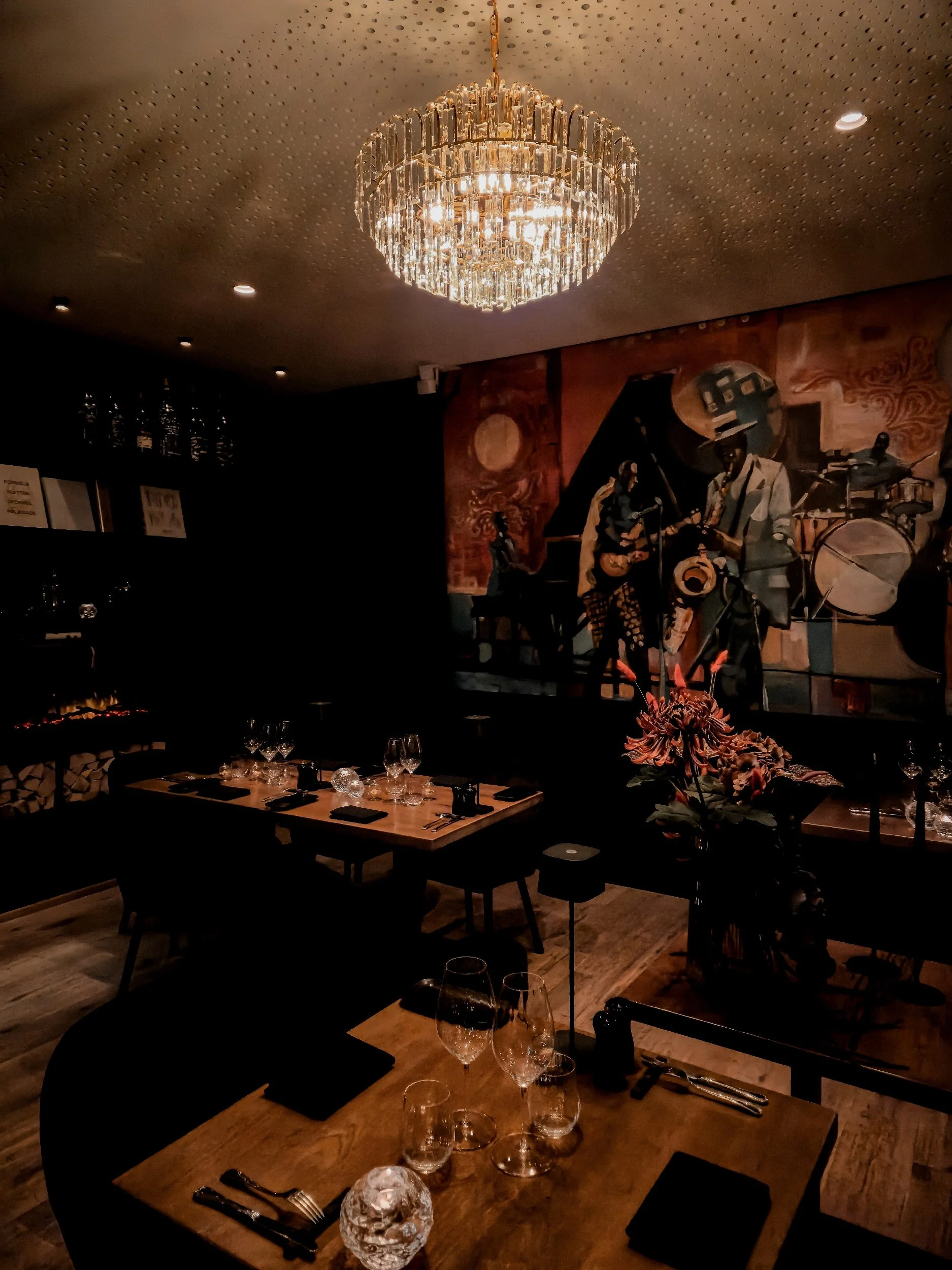 A restaurant interior with a large, ornate chandelier hanging from the ceiling, wooden tables set with wine glasses, black napkins, cutlery, and floral centerpieces, and a colorful mural of musicians playing instruments on the wall.