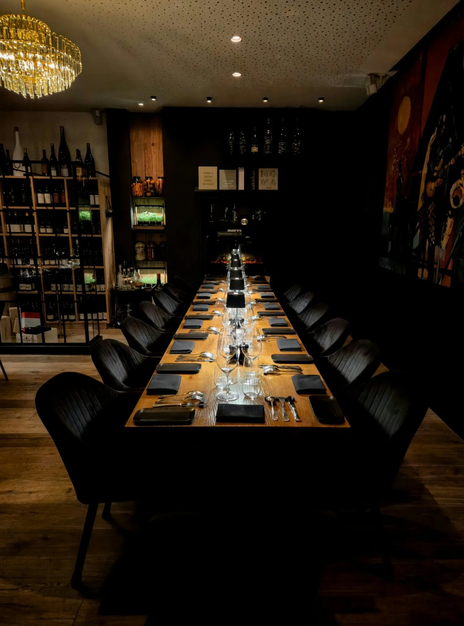 A long wooden dining table set with black napkins, glasses, and utensils in a dimly lit restaurant with wine bottles and artwork on the walls.