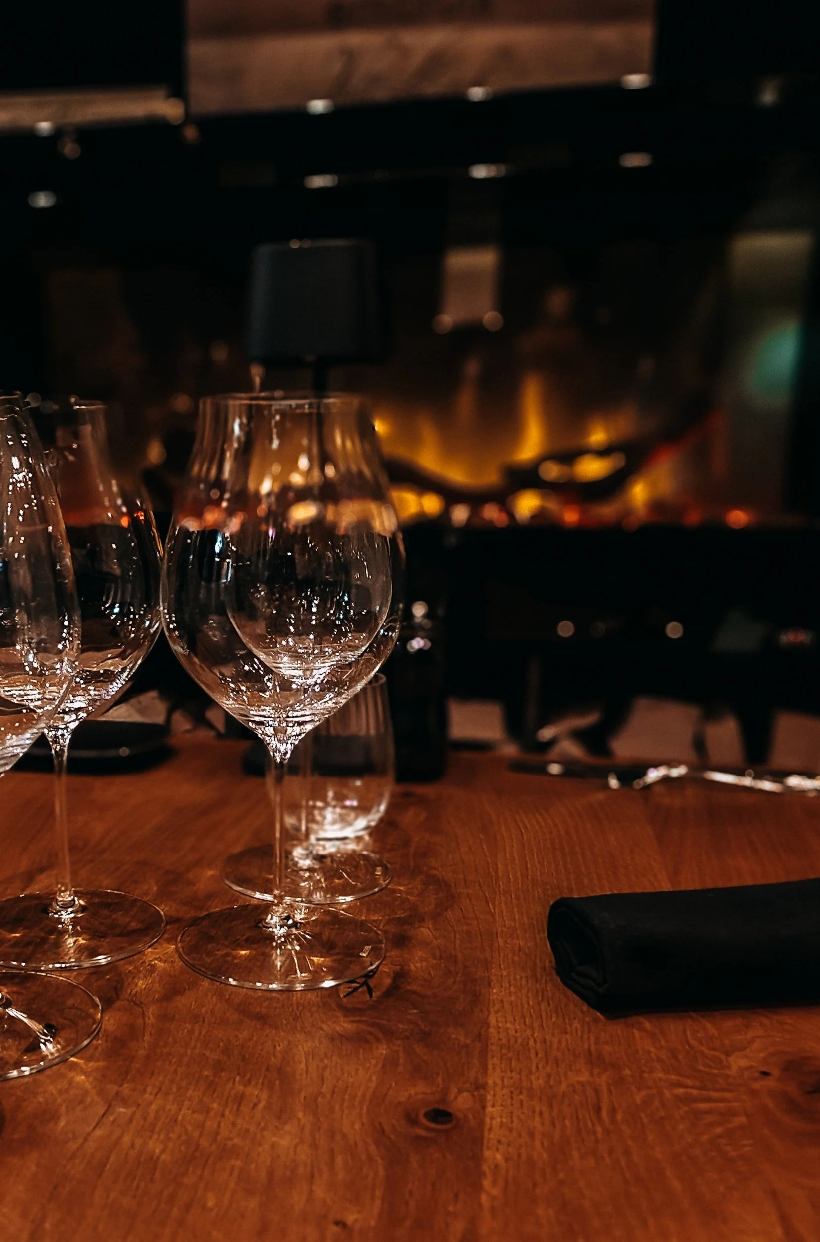 Empty wine glasses on a wooden table in a dimly lit restaurant or bar with a large fireplace or oven in the background.