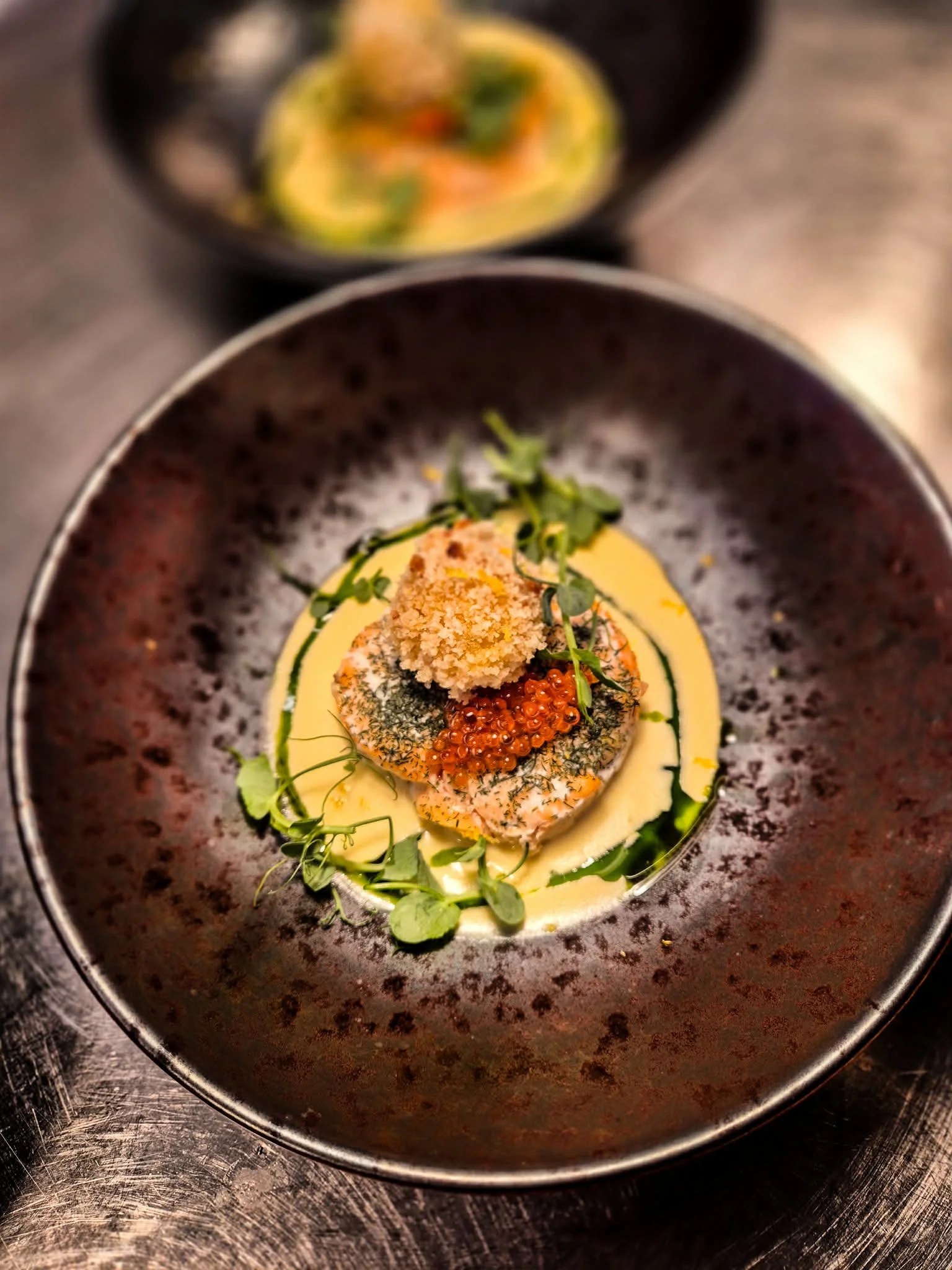 A plated dish featuring two pieces of seared fish topped with herbs, roe, and breadcrumbs, surrounded by a yellow sauce and garnished with microgreens, with another similar dish visible in the background.