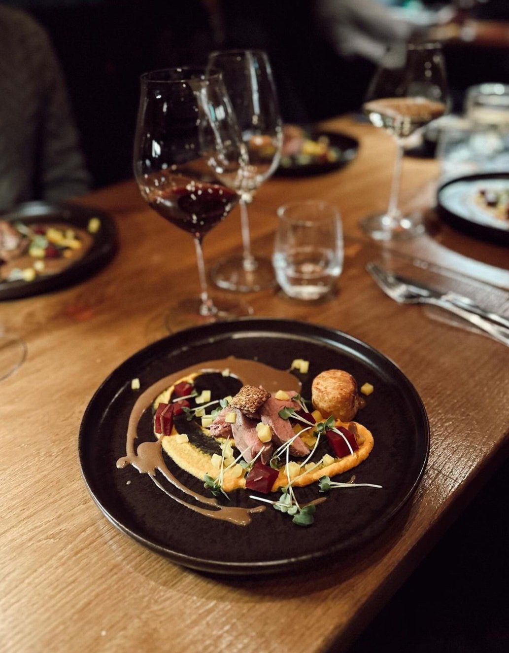 A dinner setting with a black plate featuring a gourmet dish, garnished with microgreens, accompanied by wine glasses filled with red wine, all on a wooden table with silverware and additional plates in the background.